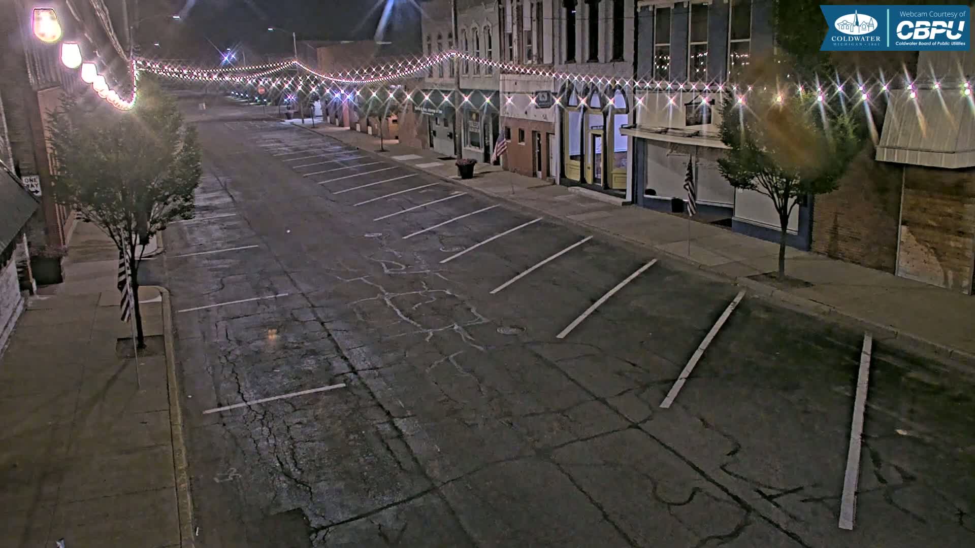 A nighttime view of a mostly empty street lined with buildings, parking spaces, and string lights.