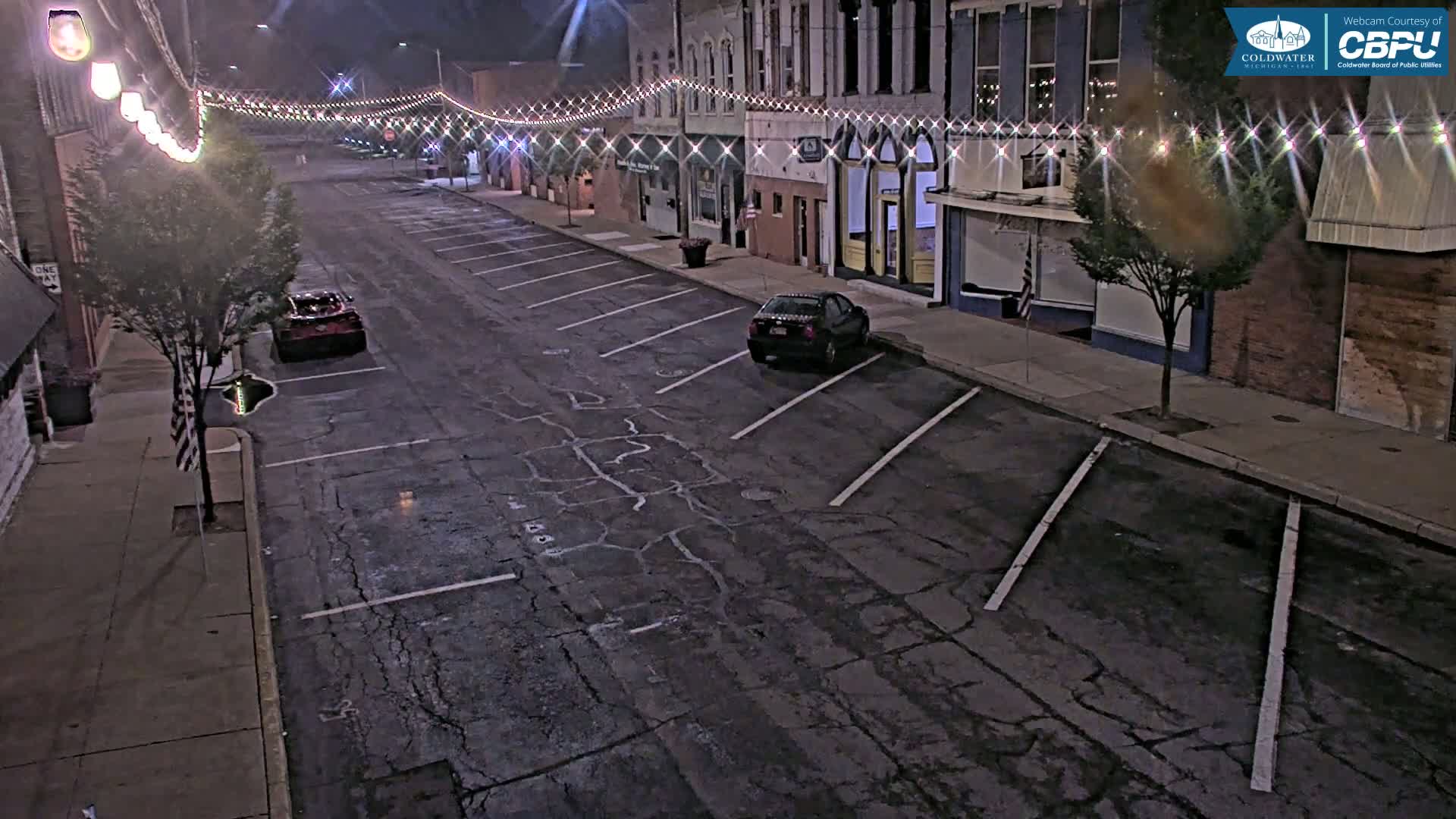 A mostly empty street at night lined with buildings decorated with string lights, and a few cars parked in the adjacent parking lot.