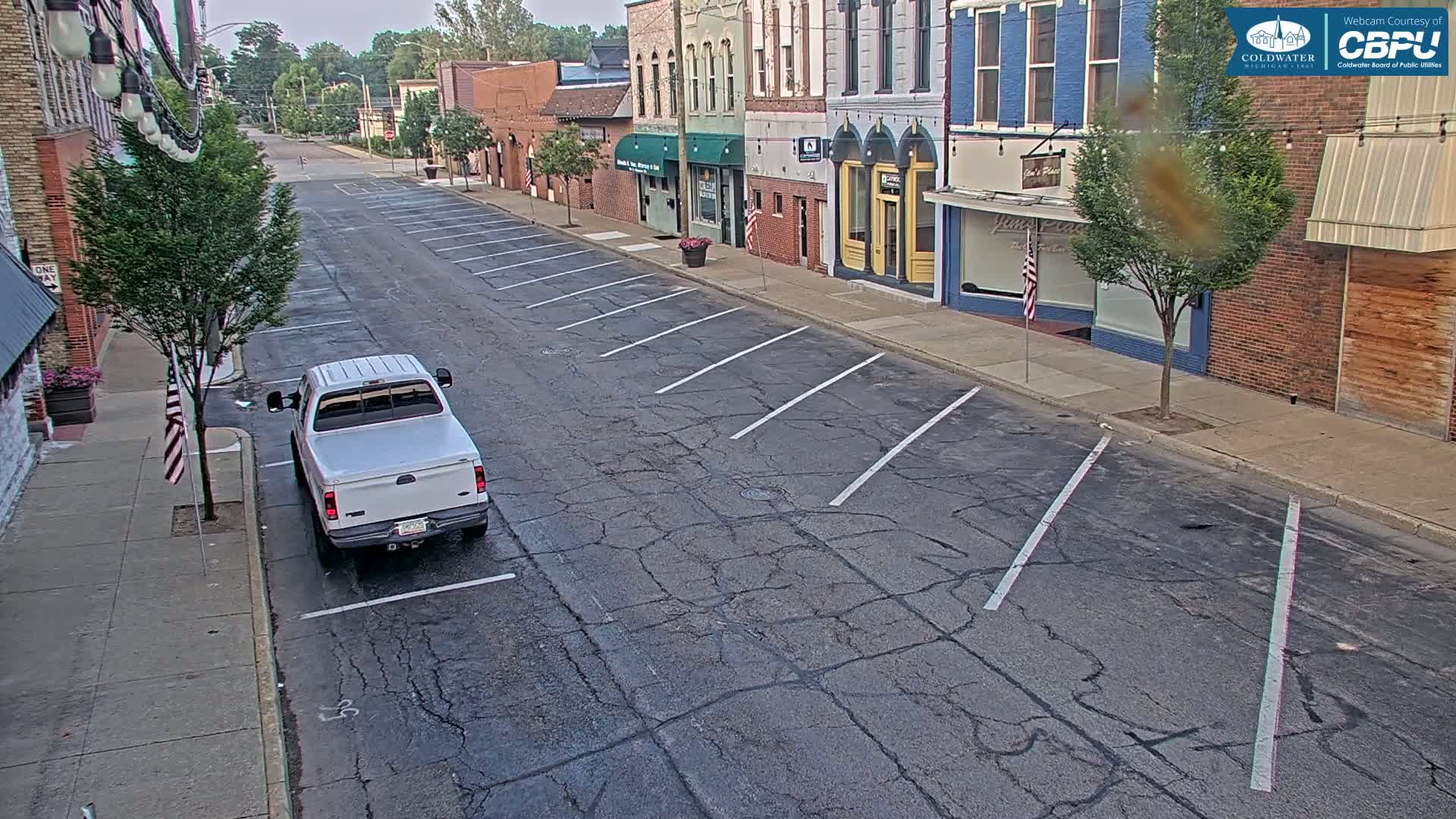 A mostly empty street with parked cars and several brick buildings lines on either side on a partly cloudy day.