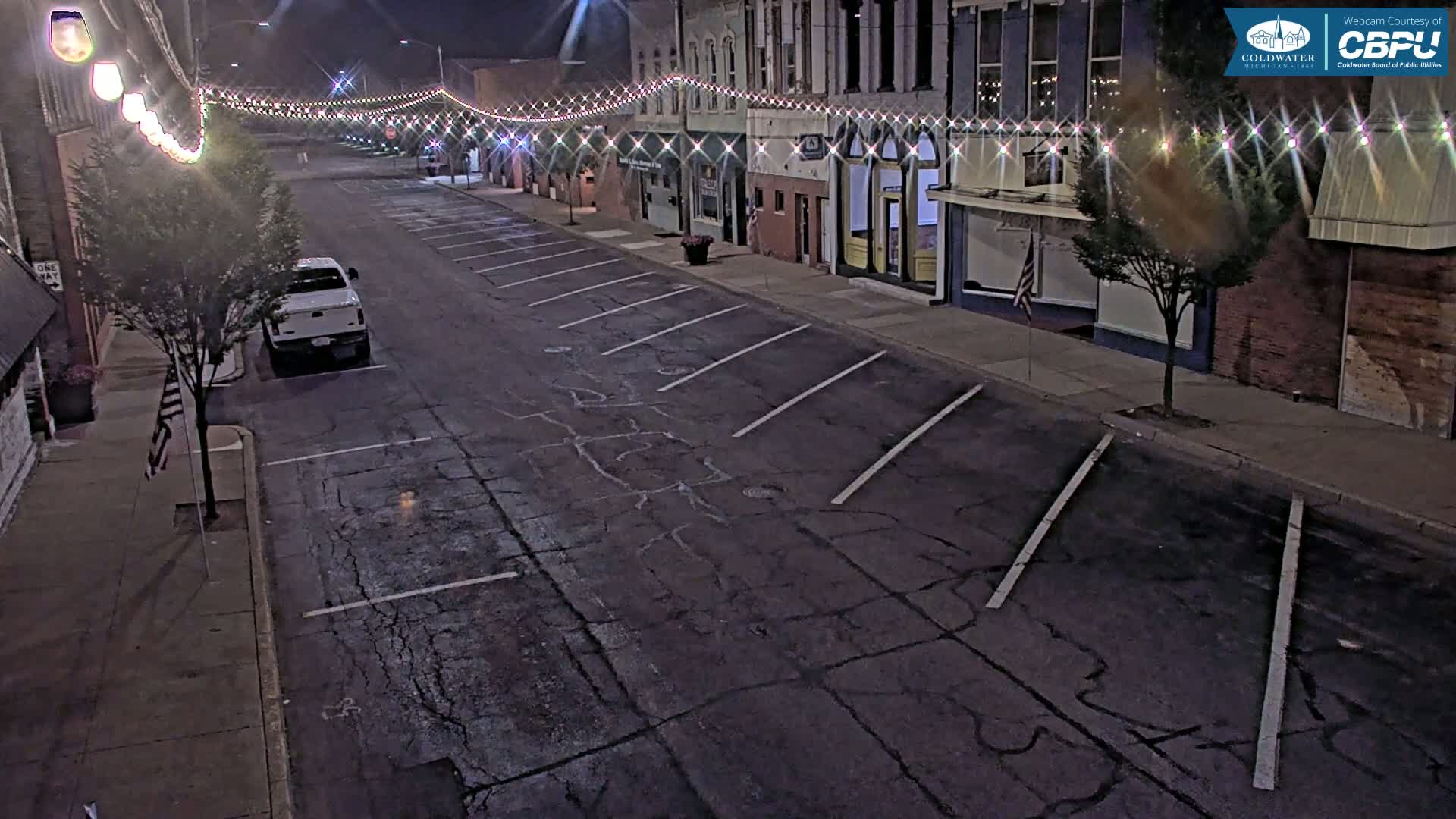 A mostly empty street at night lined with buildings decorated with string lights, has a single parked truck and cracked asphalt.