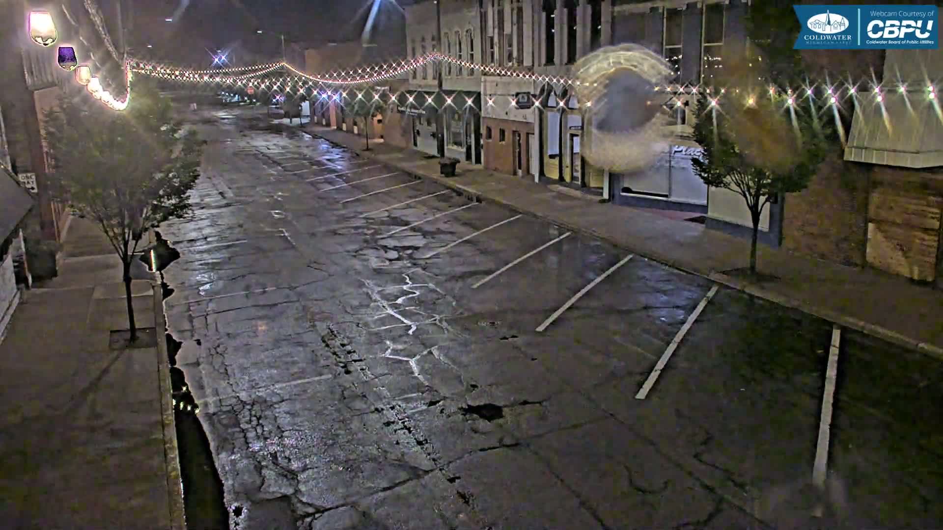 A wet, empty street at night lined with buildings decorated with string lights.