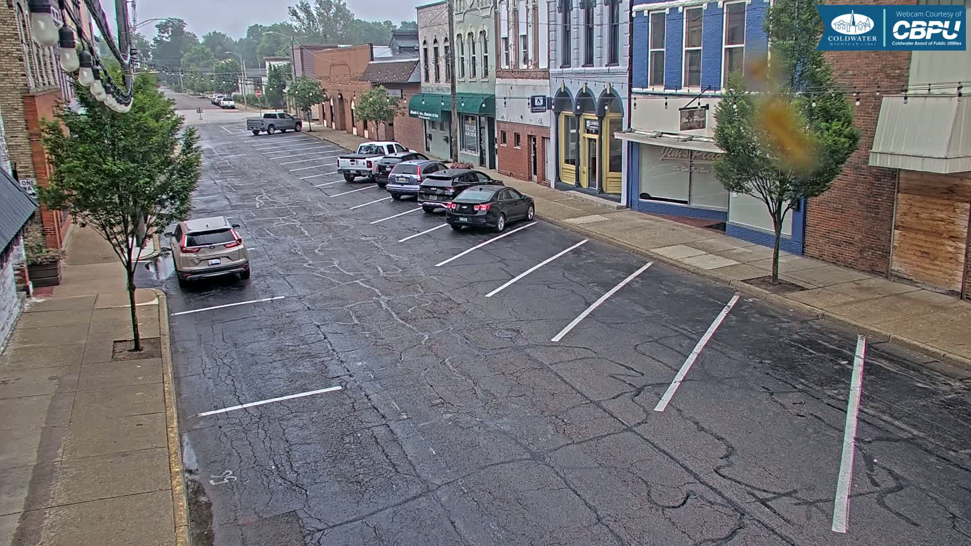 A mostly empty street with parked cars lines the sides of several brick buildings under an overcast sky.