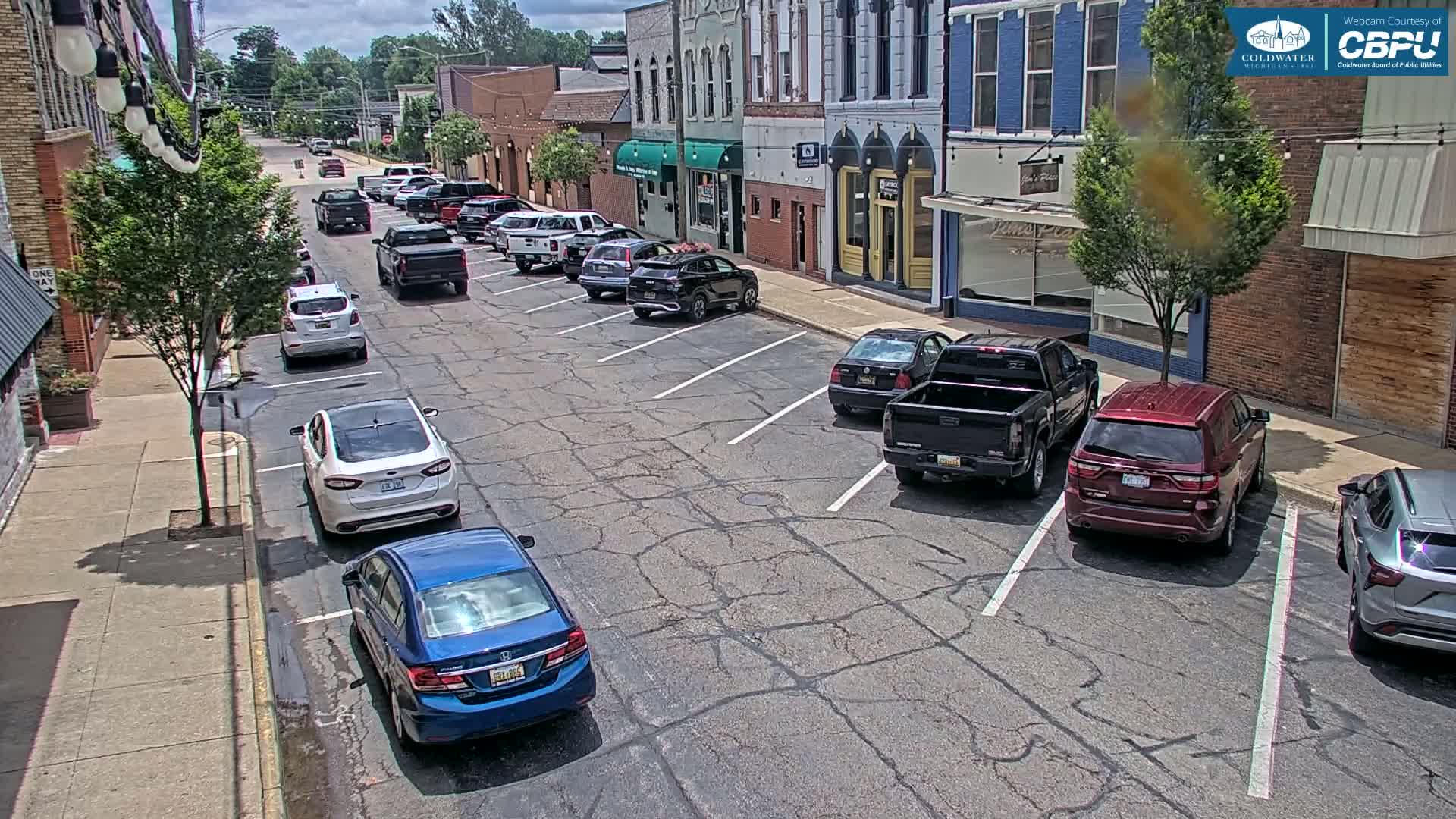 Cars are parked along the side of a street in front of brick buildings on a partly sunny day.
