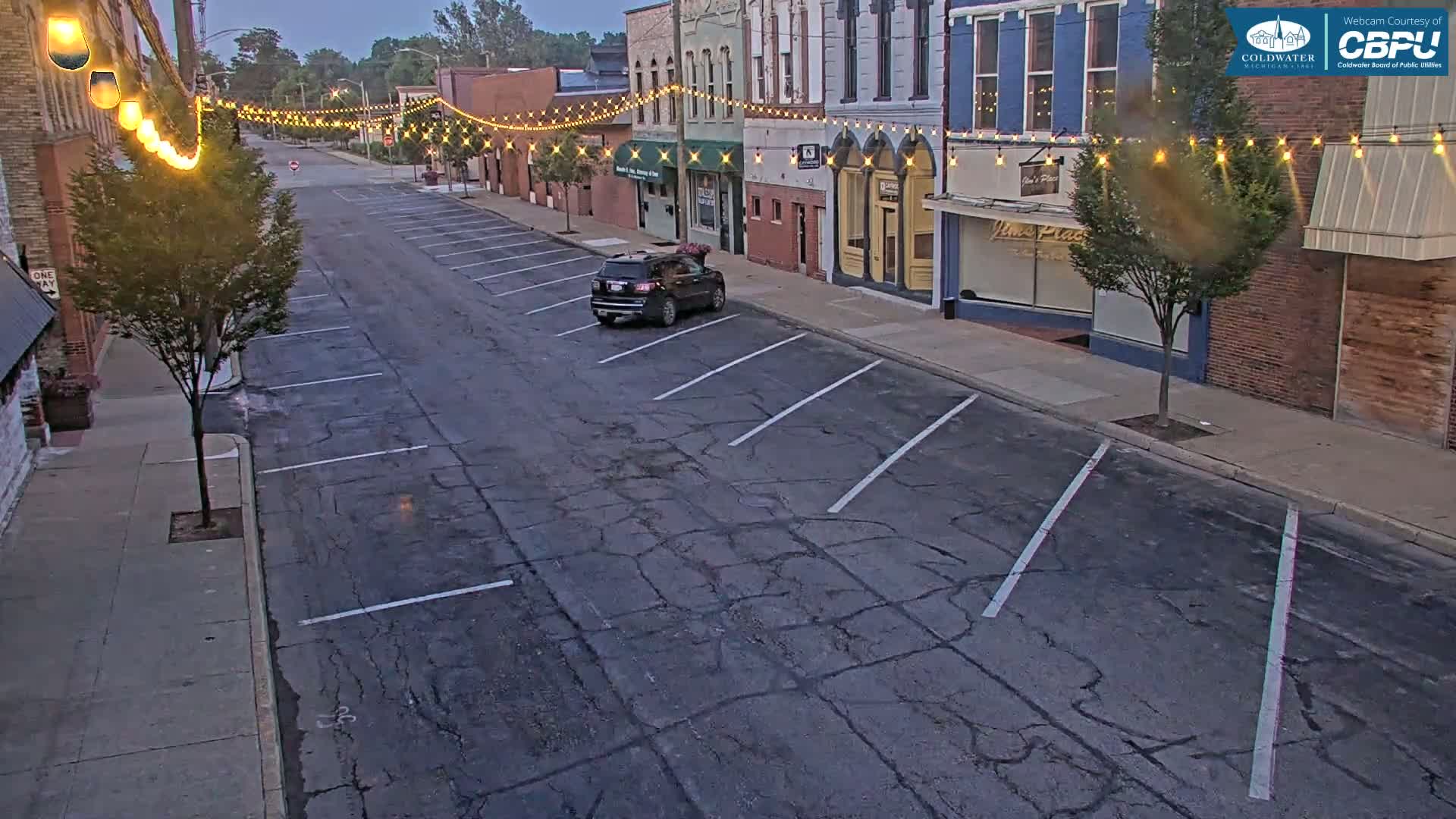 A mostly empty street with parked cars, lined with brick buildings decorated with string lights under a twilight sky.