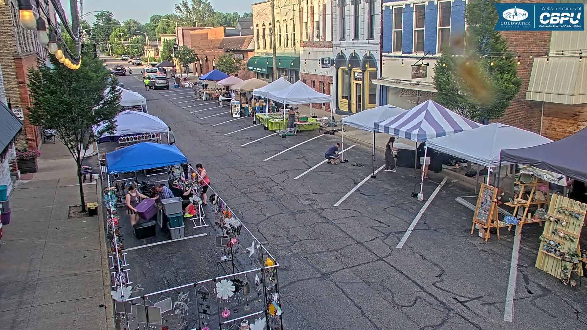 A street market with numerous vendor tents is set up in a town's parking lot on a partly sunny day.