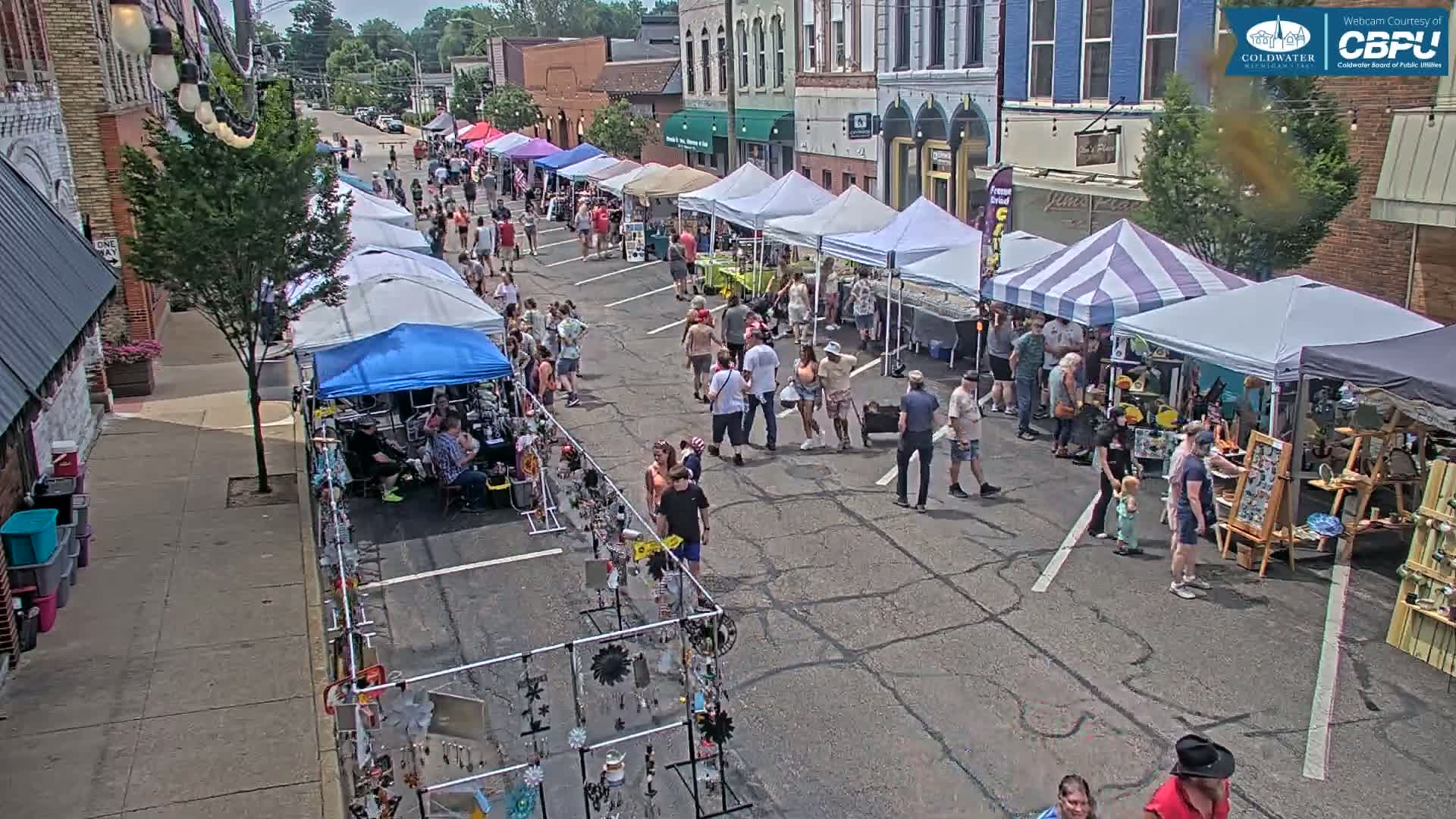 A street market with numerous vendors under canopies is bustling with people on a sunny day.