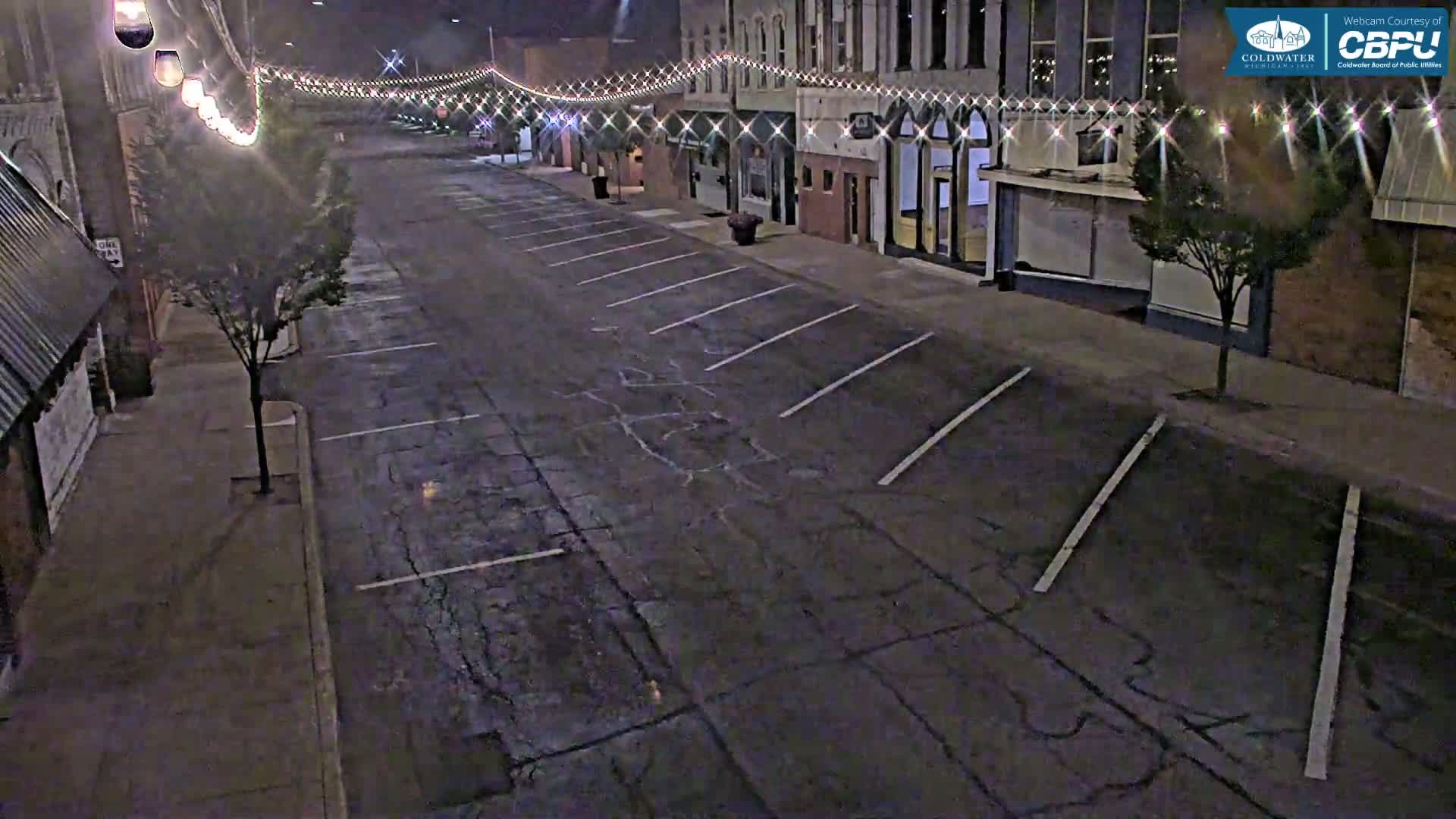 A mostly empty street at night lined with buildings, illuminated by string lights, and featuring parking spaces on either side.