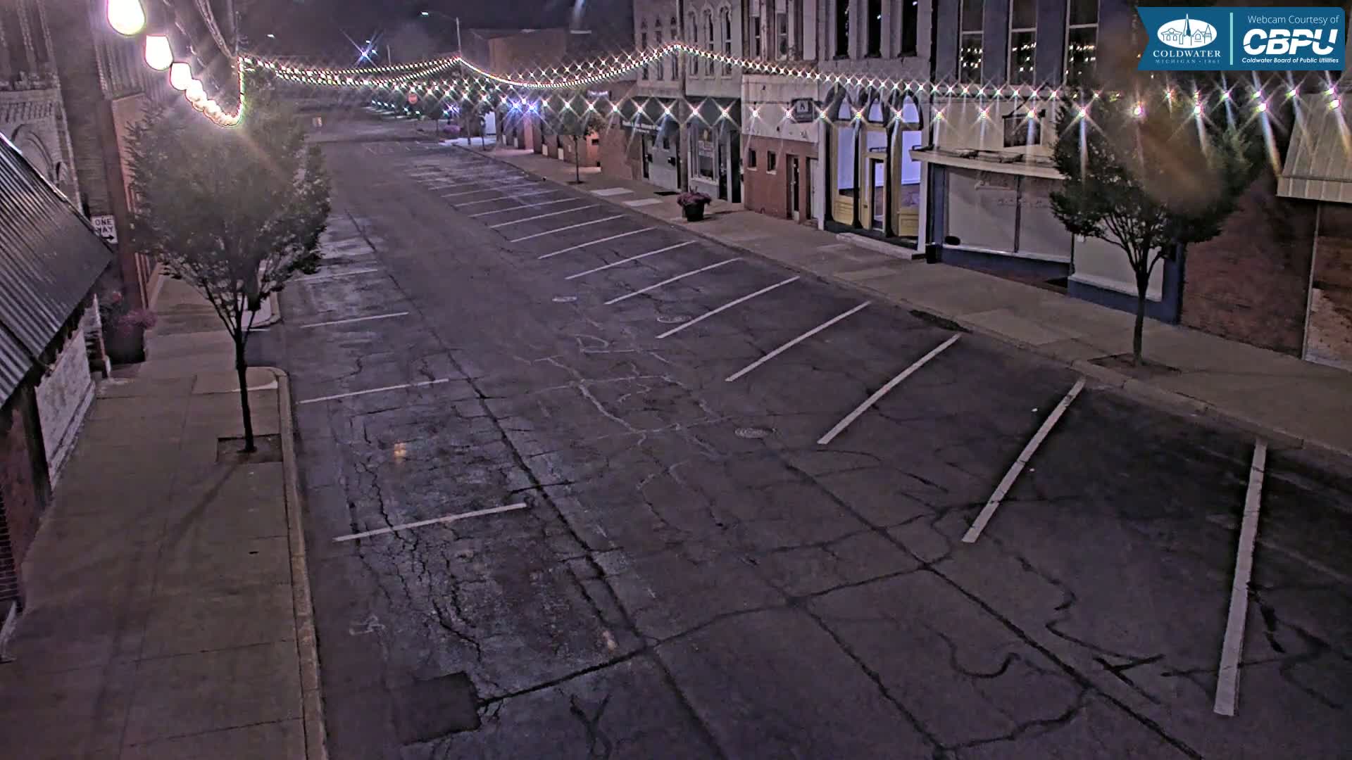 A nighttime street scene shows a mostly empty parking lot next to a row of buildings decorated with string lights.