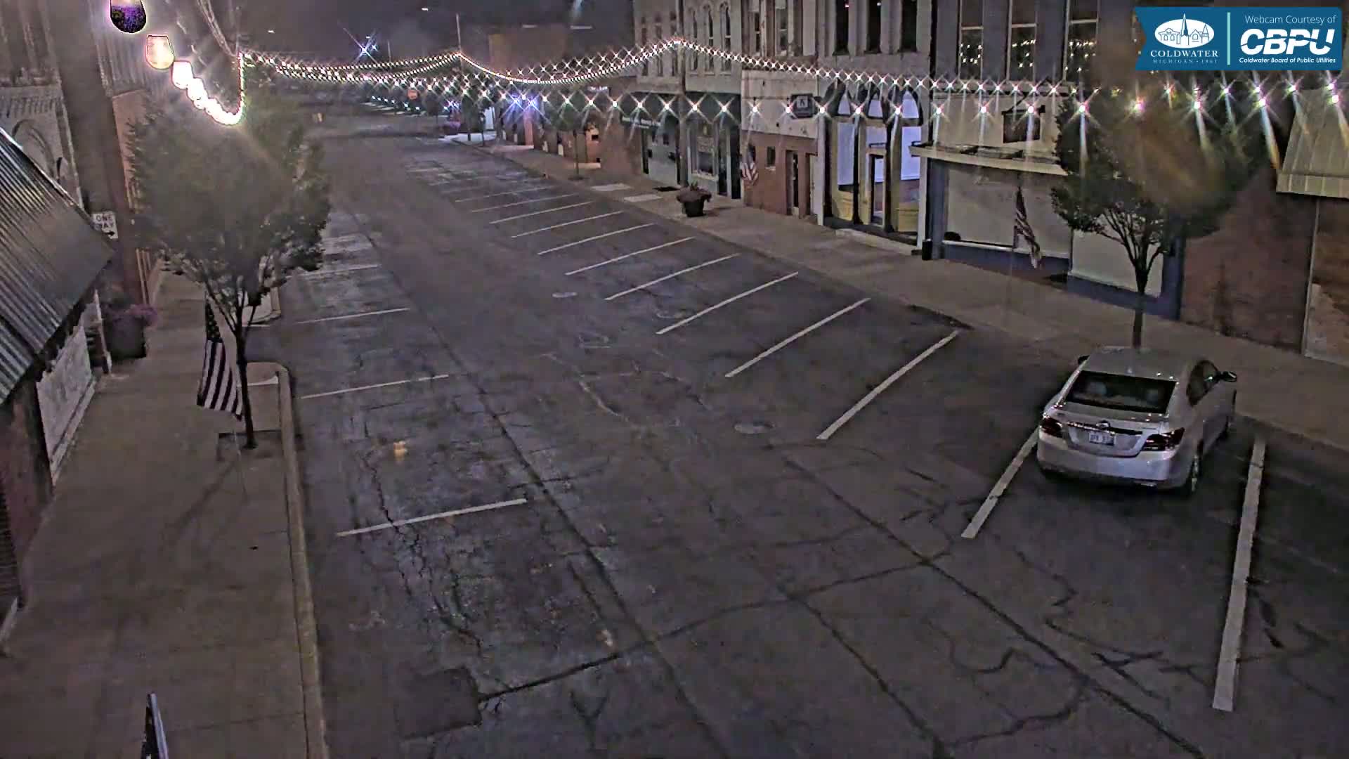 A mostly empty street at night, lined with buildings and parking spaces, is illuminated by streetlights and string lights.