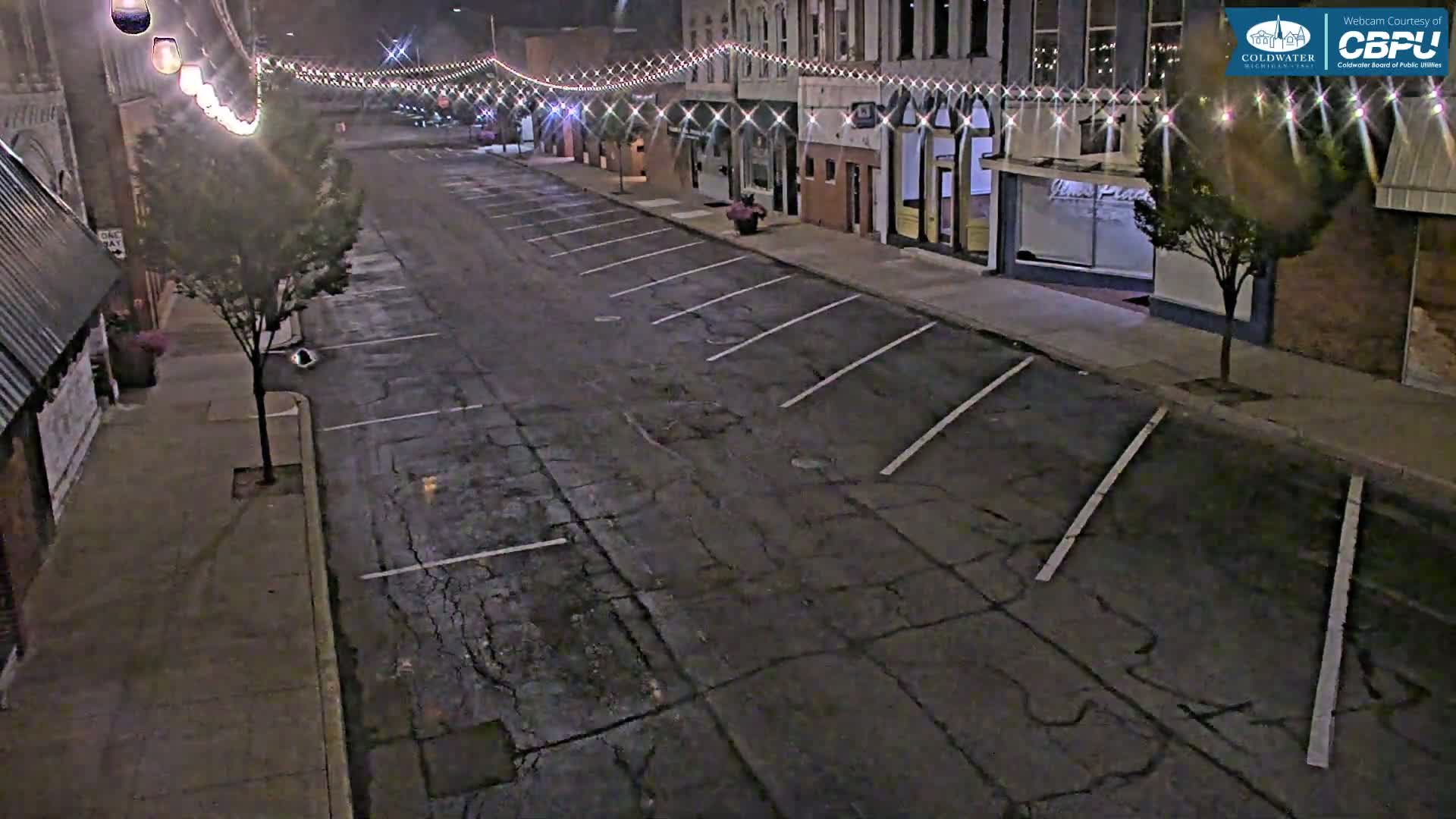 A nighttime street scene shows a nearly empty parking lot in front of buildings decorated with string lights.