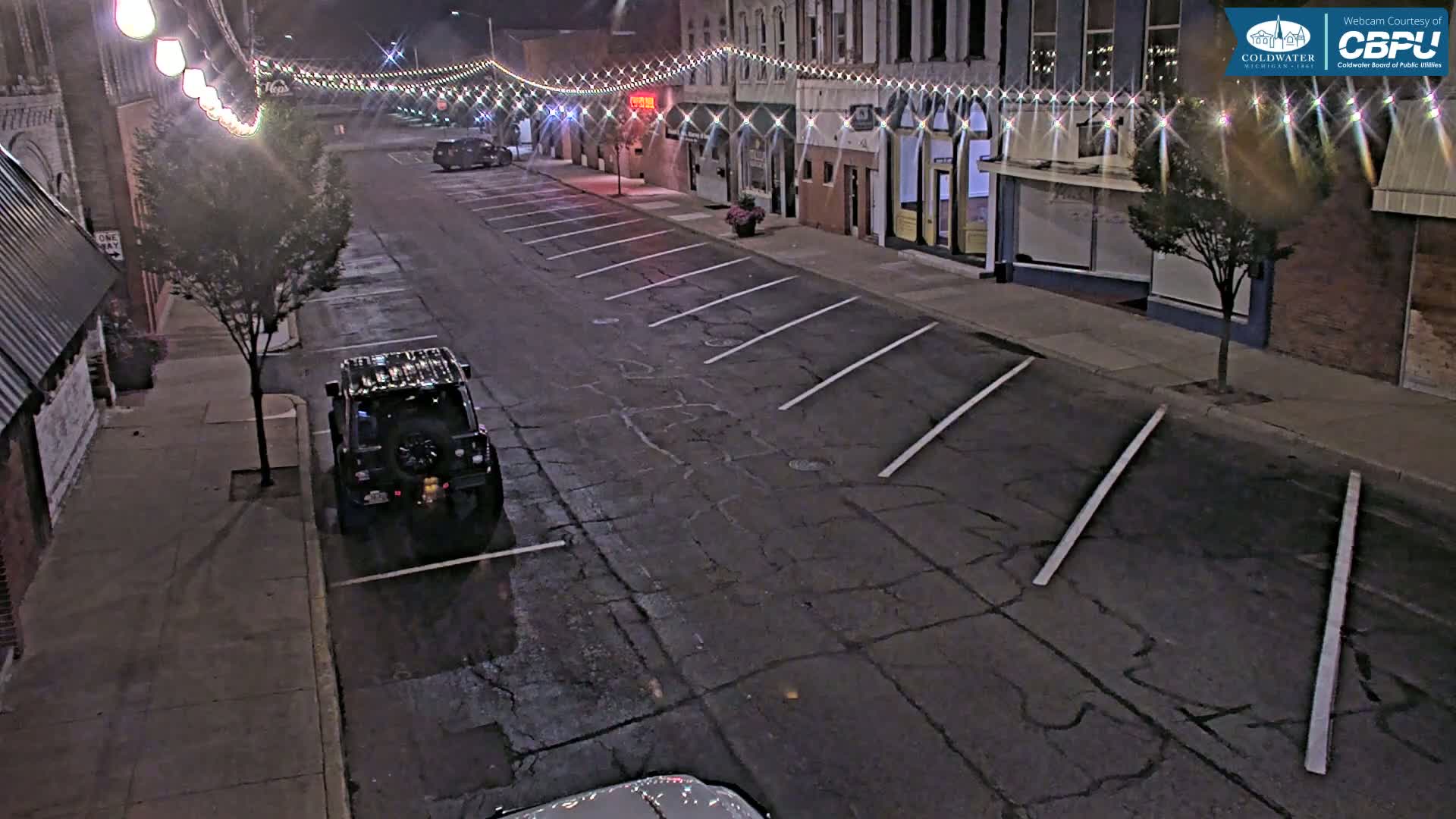 A nighttime street scene shows a nearly empty parking lot in front of buildings decorated with string lights.