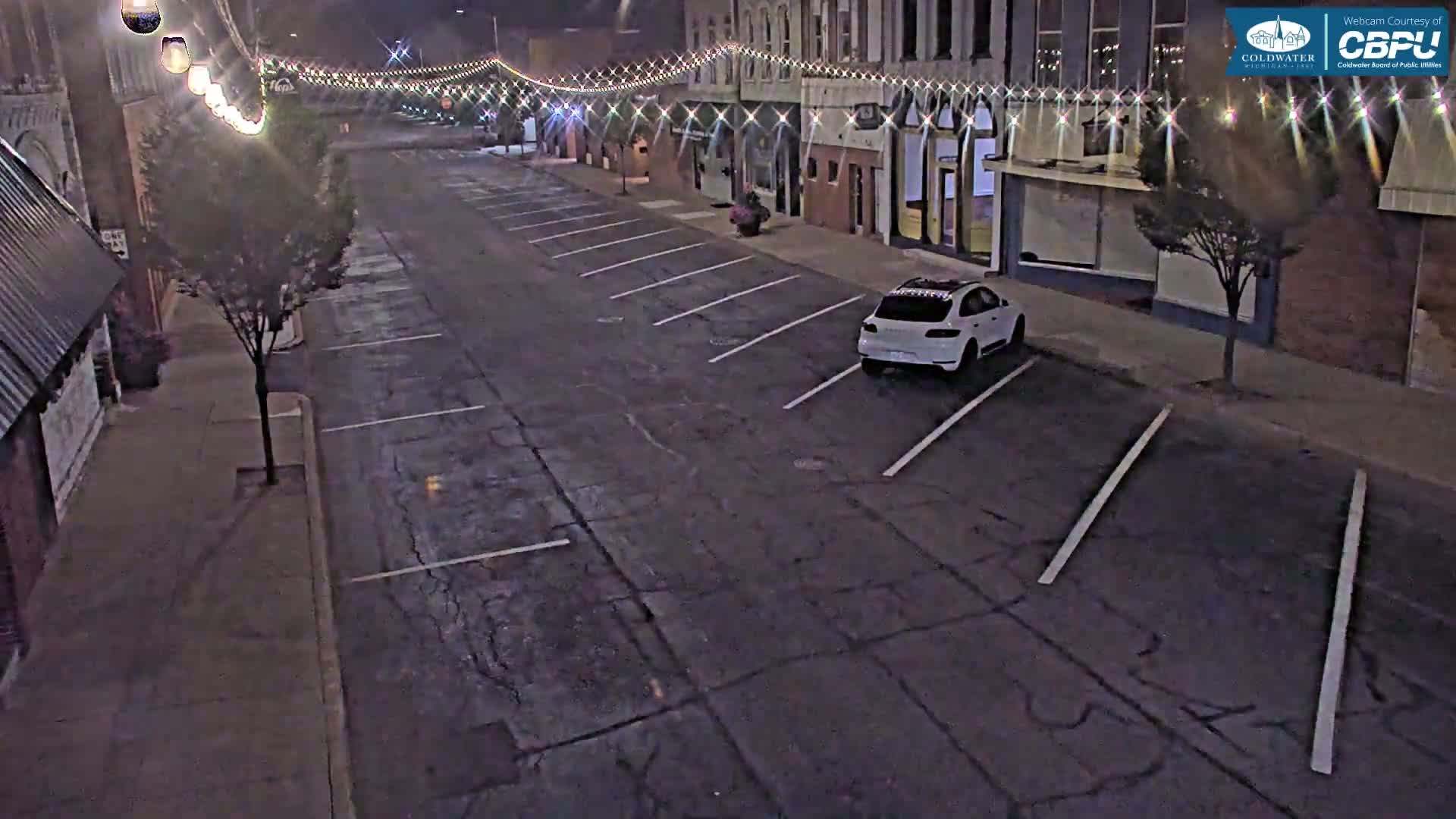 A white car is parked in an empty parking lot at night, next to a street lined with buildings decorated with string lights.