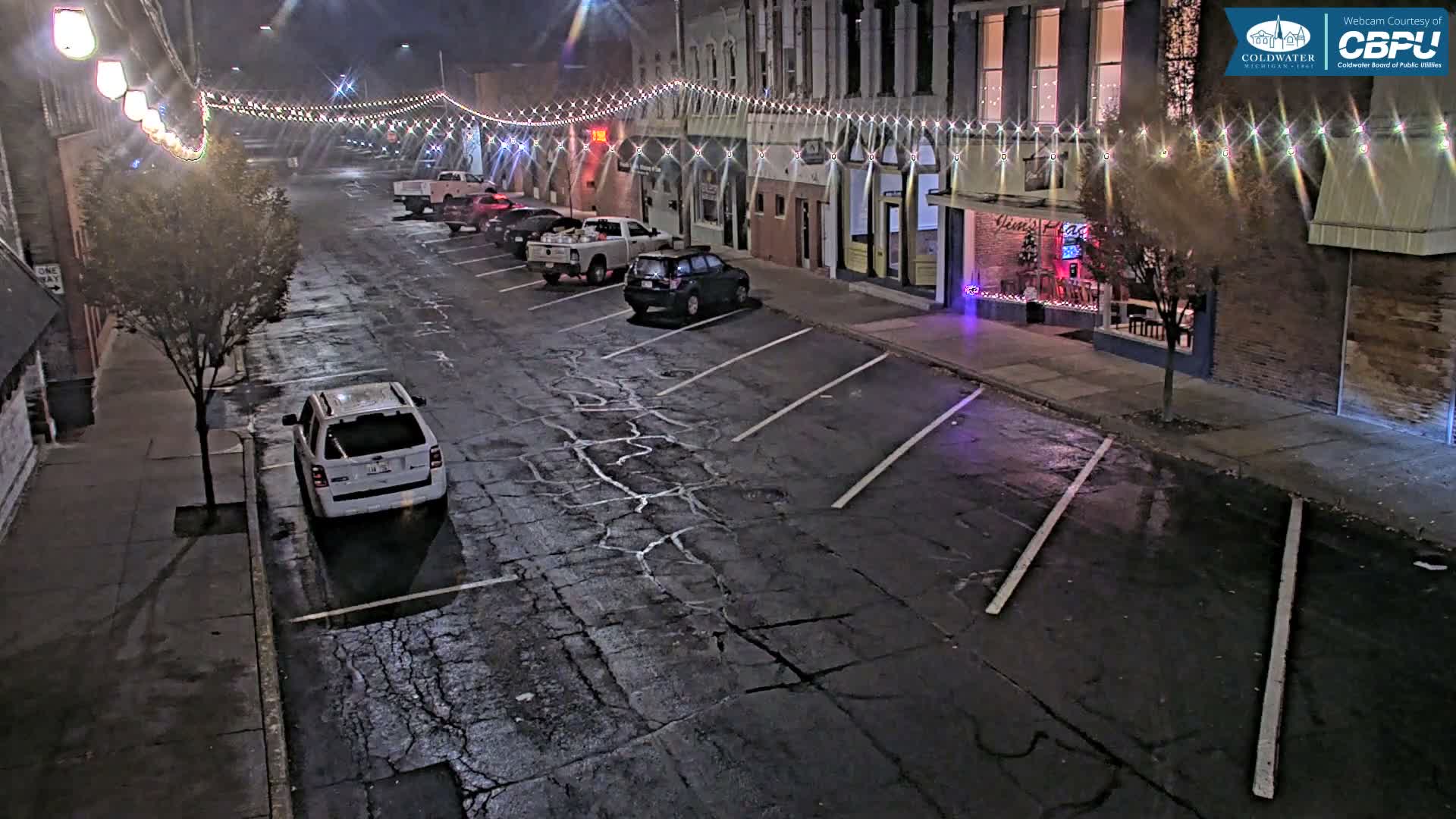 A white car is parked in an empty parking lot at night, next to a street lined with buildings decorated with string lights.