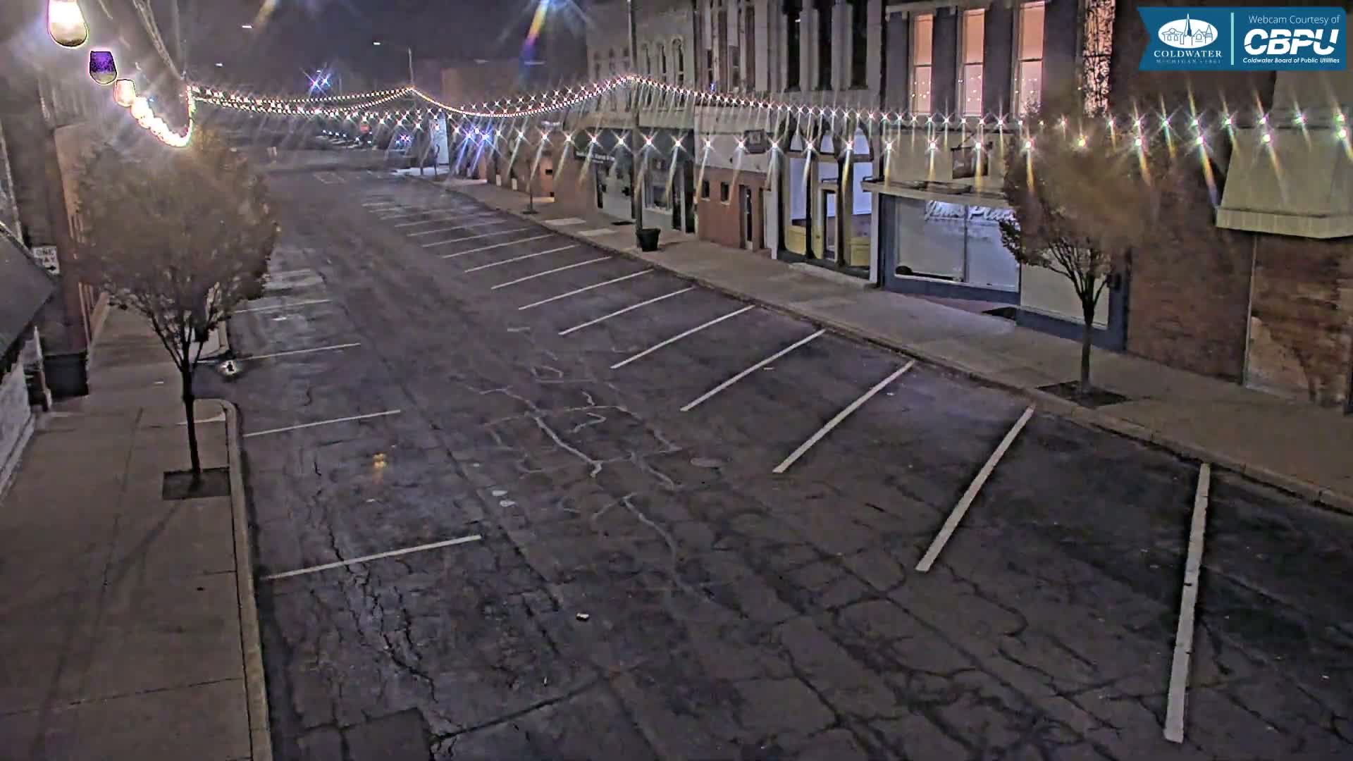 An empty, cracked asphalt street with diagonal parking lines is illuminated by overhead string lights, flanked by buildings and a few small trees under a clear night sky.