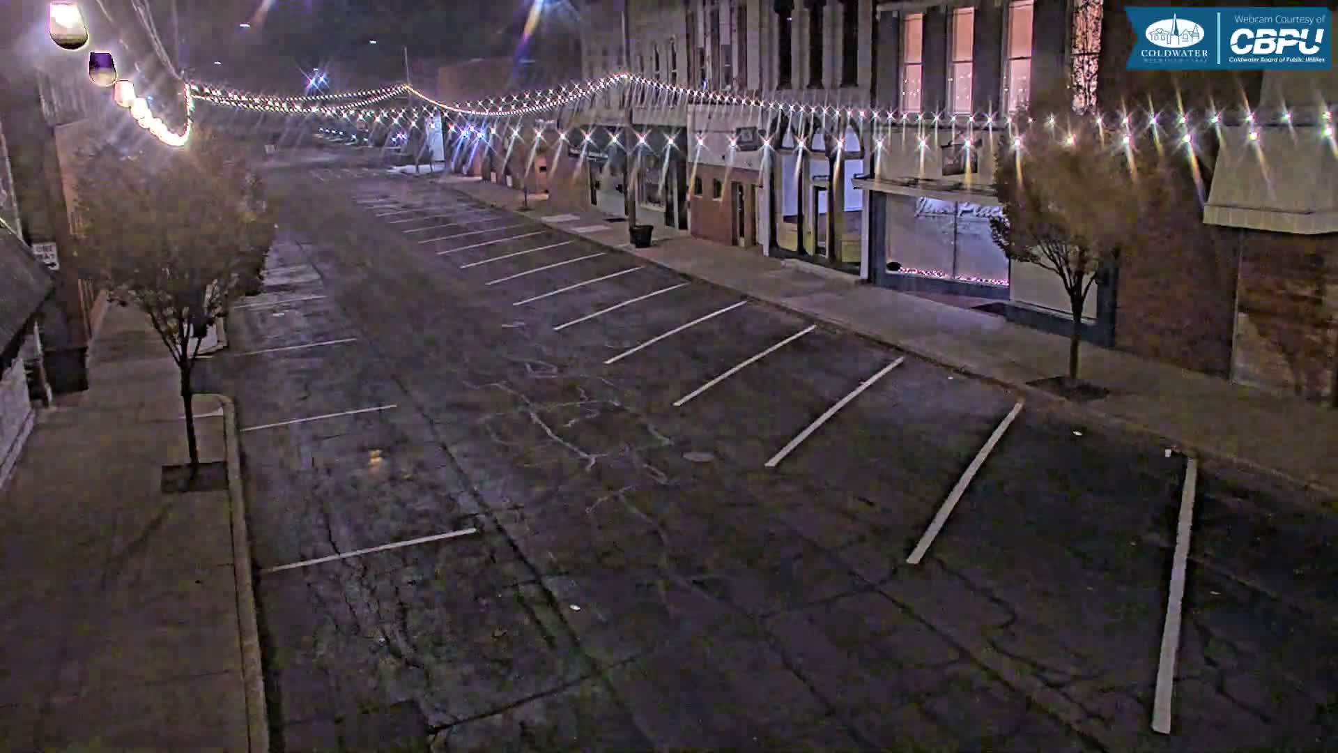 An empty, wet street at night is illuminated by festive string lights strung above a row of brick buildings and several trees on the sidewalks.