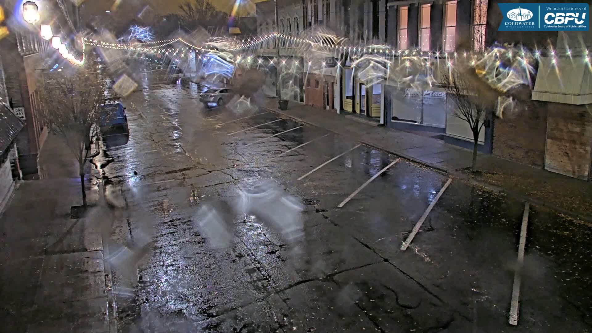 A blurry, rain-streaked nighttime webcam view captures a wet street with parked cars, reflective cracked pavement, string lights strung across buildings, and a few bare trees.