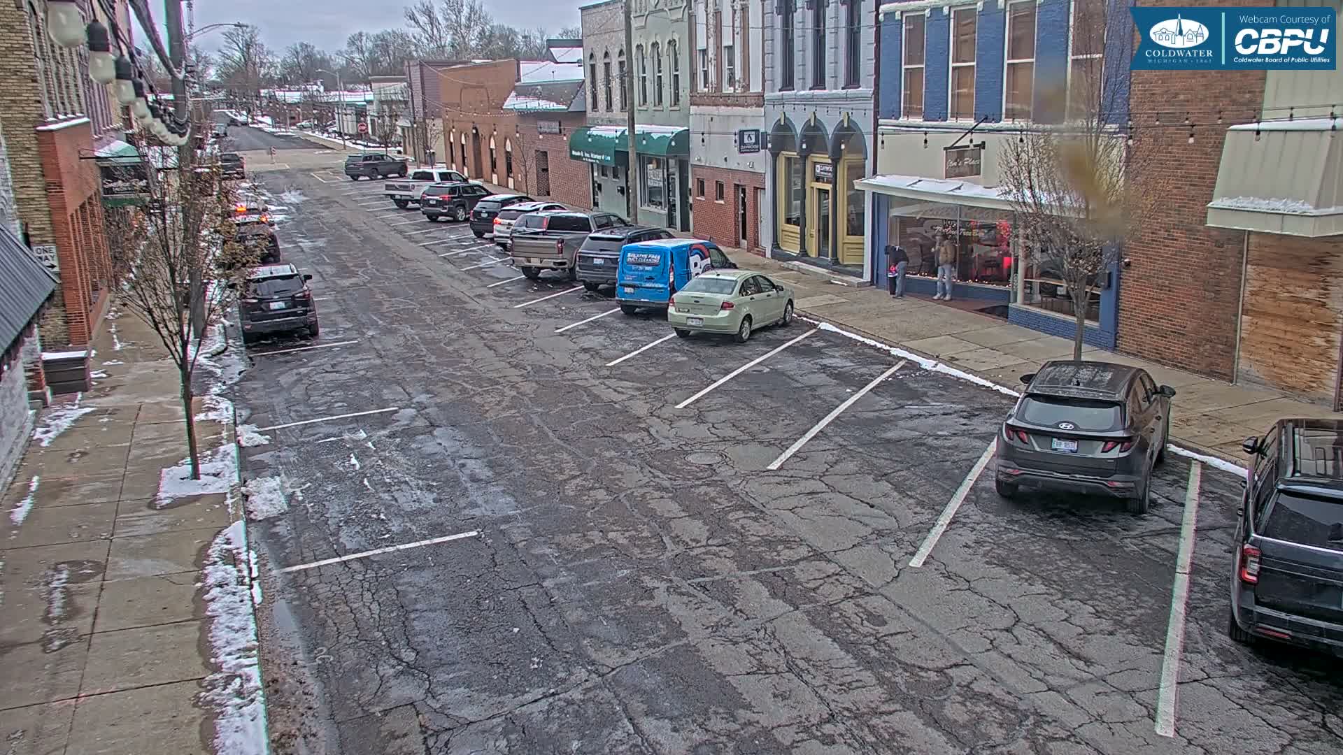 A street in a town is visible, lined with multi-story buildings and numerous parked cars, with patches of melting snow on the sidewalks and cracked pavement under an overcast sky.