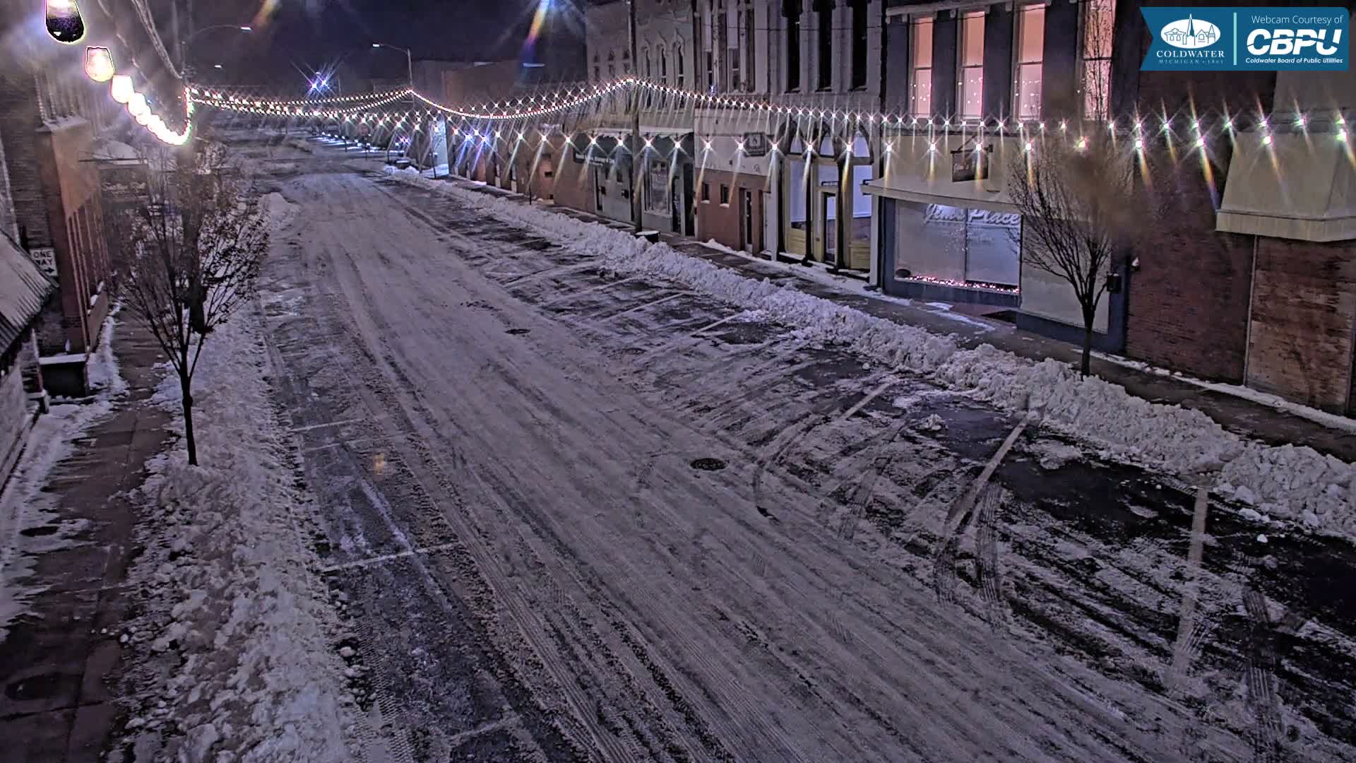 A quiet, snow-covered street at night is illuminated by festive overhead string lights, with buildings lining both sides and tire tracks visible on the cold pavement.