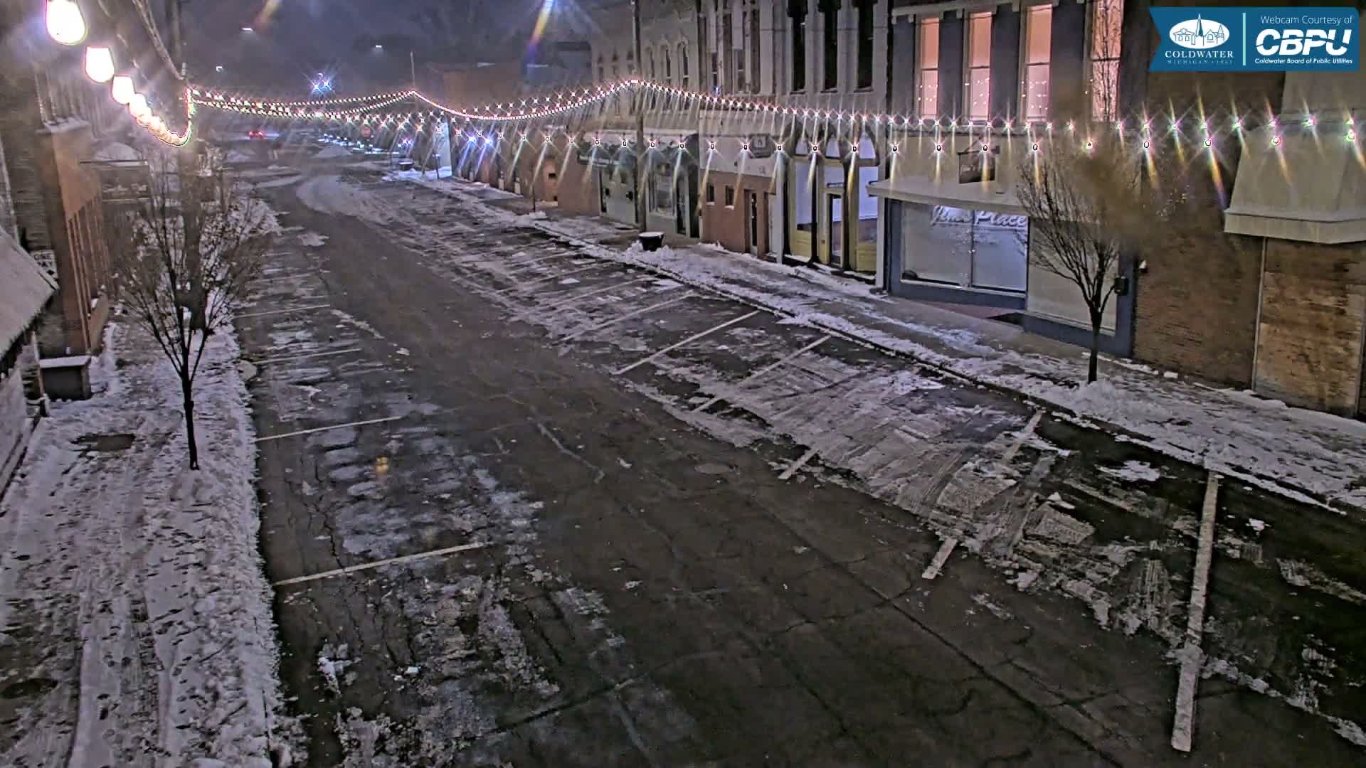 An empty downtown street, lined with buildings and adorned with illuminated string lights overhead, is partially covered in snow and slush under a dark night sky.
