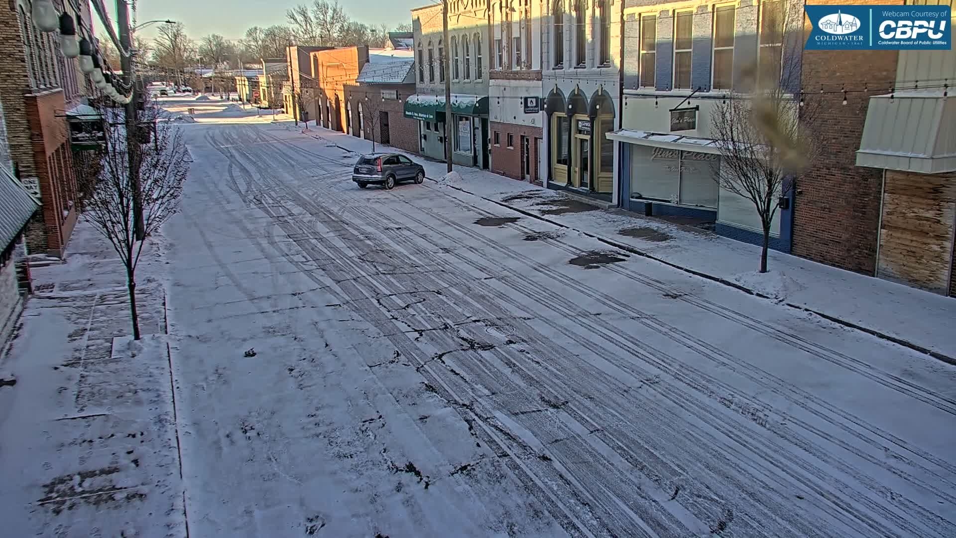 An empty downtown street, lined with buildings and adorned with illuminated string lights overhead, is partially covered in snow and slush under a dark night sky.