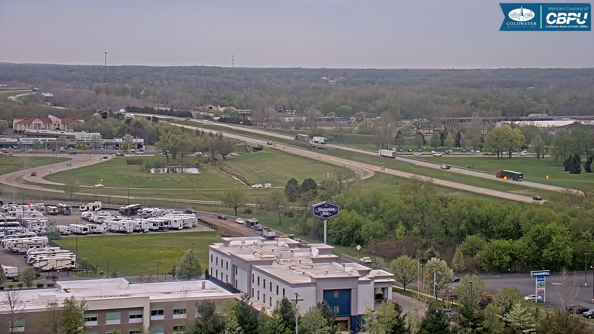 A highway with moderate traffic runs through a green landscape dotted with buildings, including a hotel and a large RV park, under a mostly clear sky.