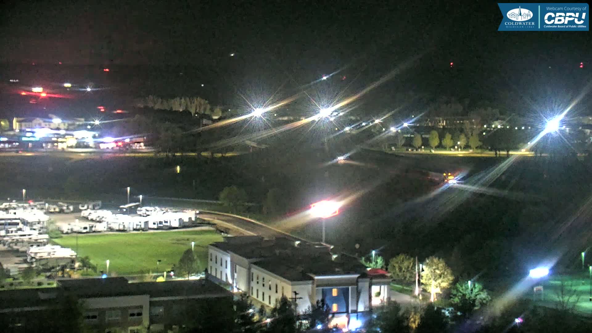 A nighttime, high-angle view of a landscape featuring a parking lot filled with RVs, a multi-story building, and distant lights under a clear, dark sky.