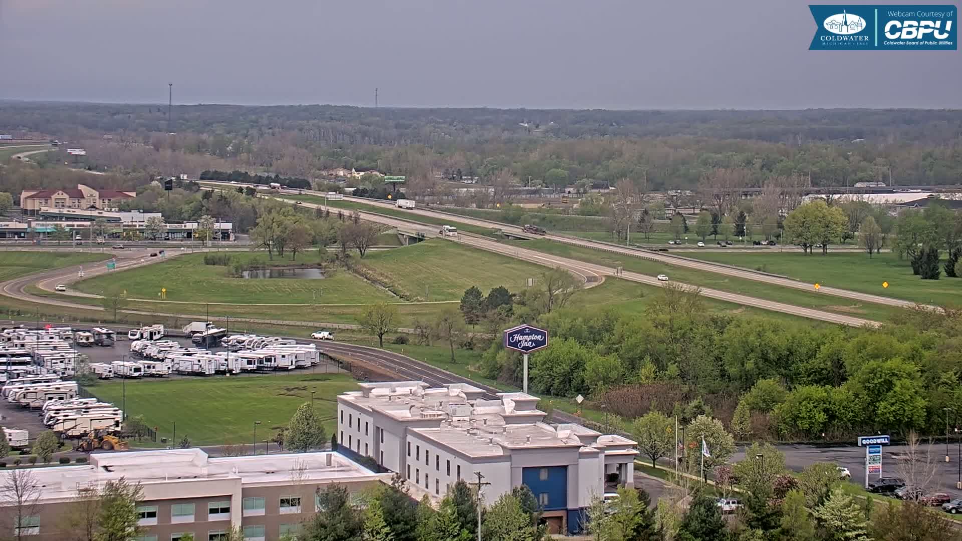 A high-angle view shows a highway, a parking lot full of RVs, a Hampton Inn, and surrounding green spaces under an overcast sky.