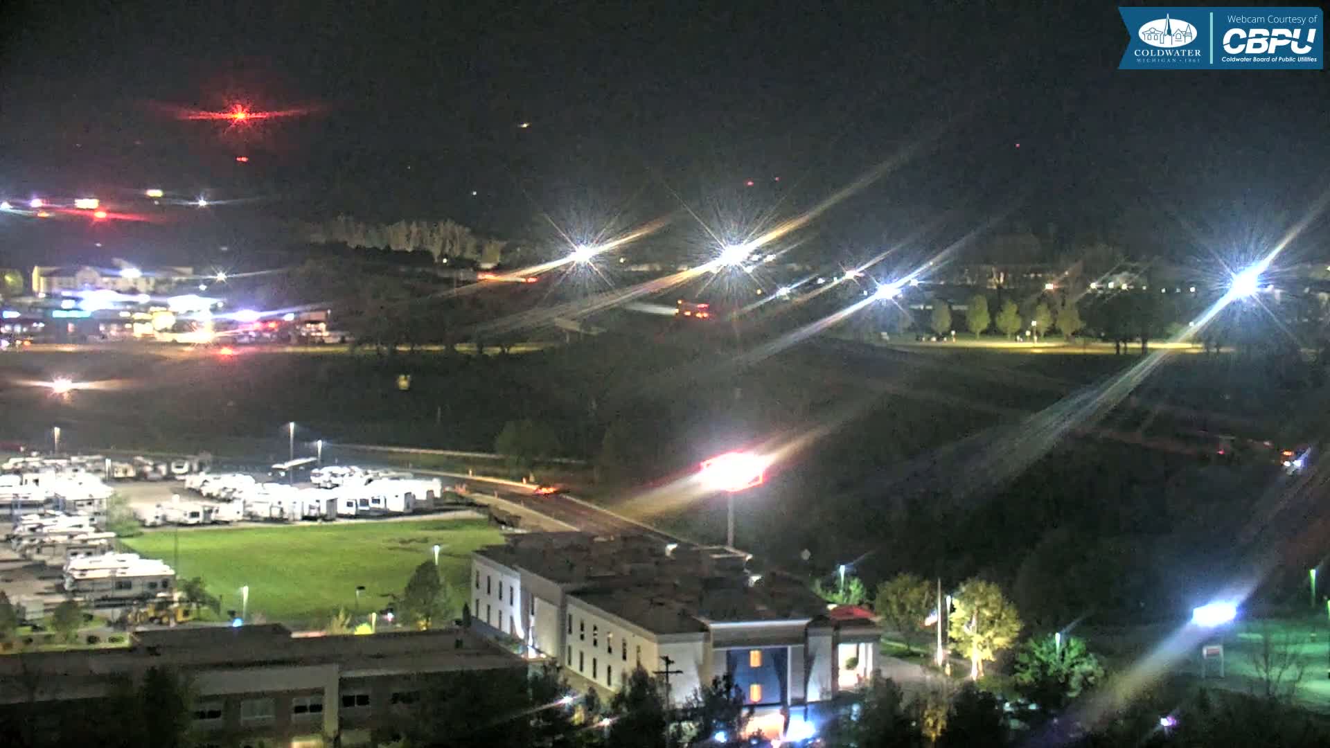 A nighttime aerial view shows a highway with cars driving on it, a parking lot full of RVs, a large building, and a field under a clear, dark sky.