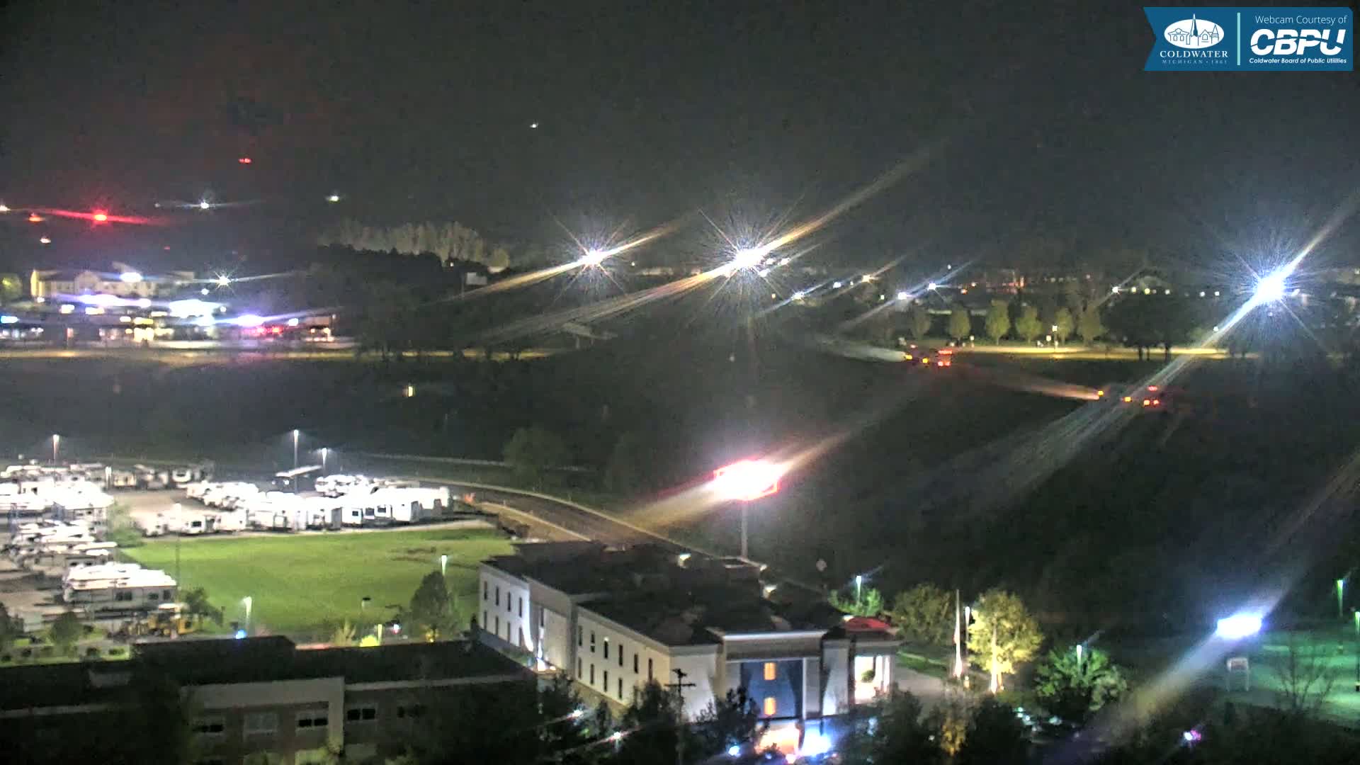 A nighttime aerial view shows a highway with cars driving, a large parking lot filled with RVs, a hotel, and other buildings, under clear skies.
