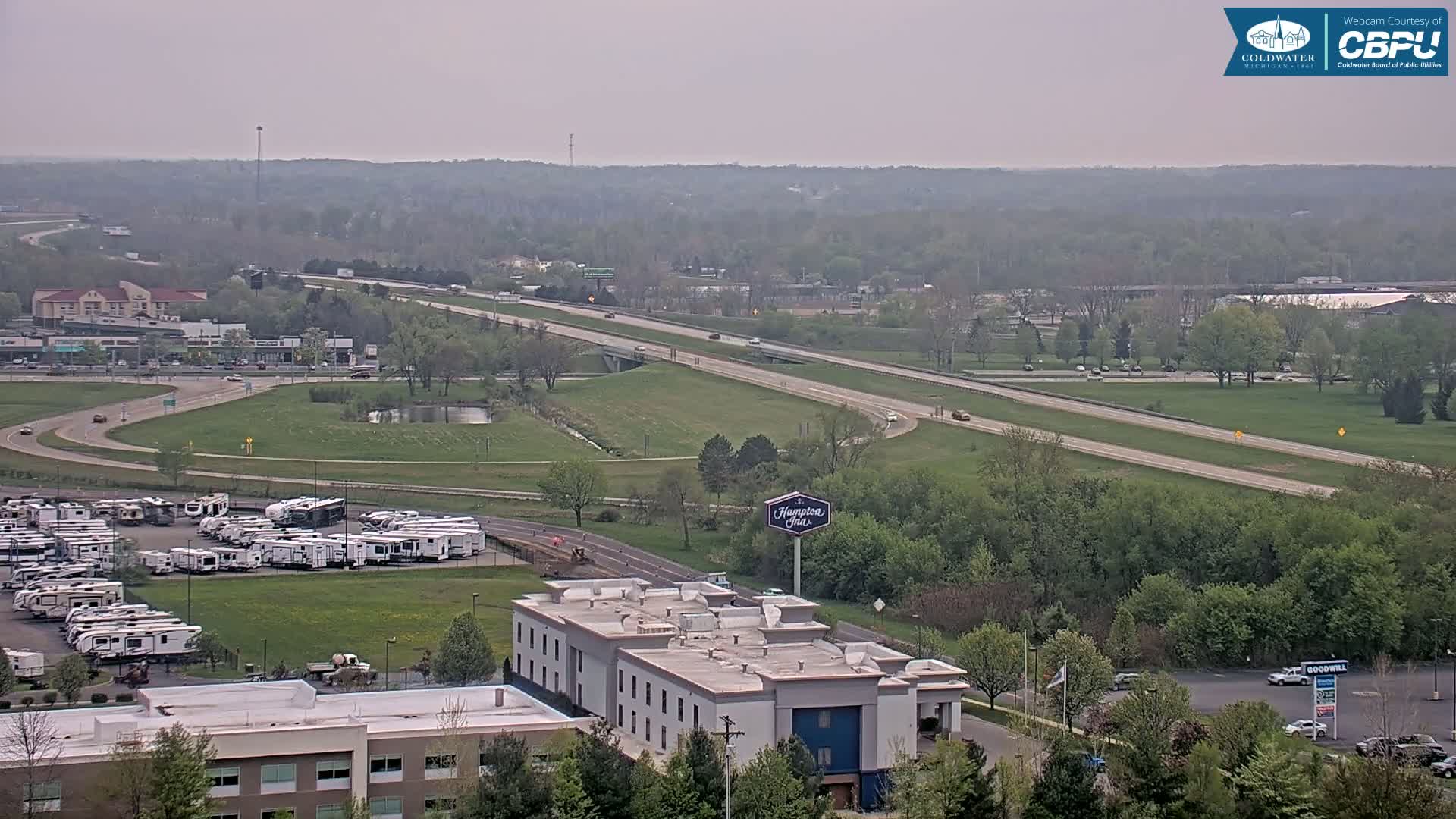 A hazy, overcast day reveals a highway, a large parking lot full of RVs, a Hampton Inn, and a Goodwill store situated amidst green, leafy trees and grassy areas.