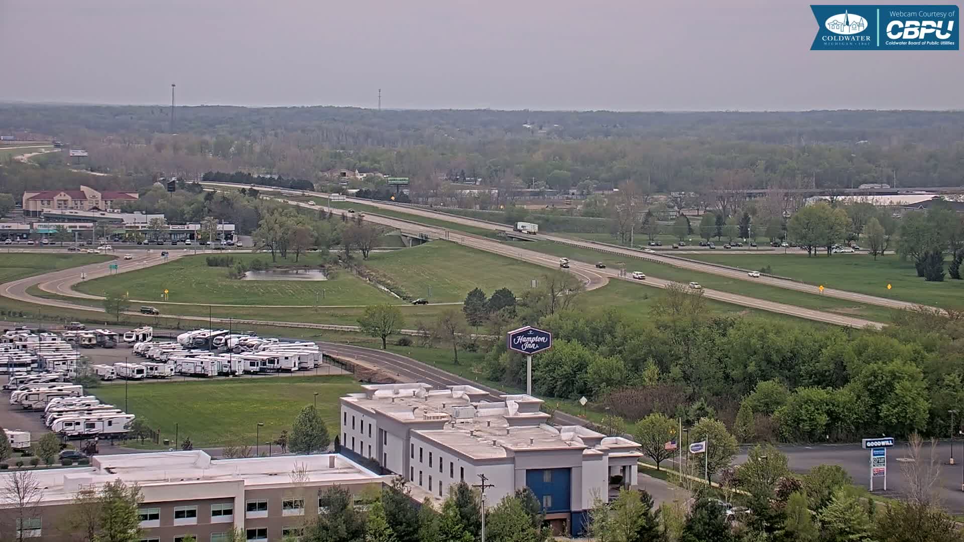 A highway with moderate traffic runs through a green landscape dotted with buildings, including a hotel and a large RV park, under a mostly cloudy sky.