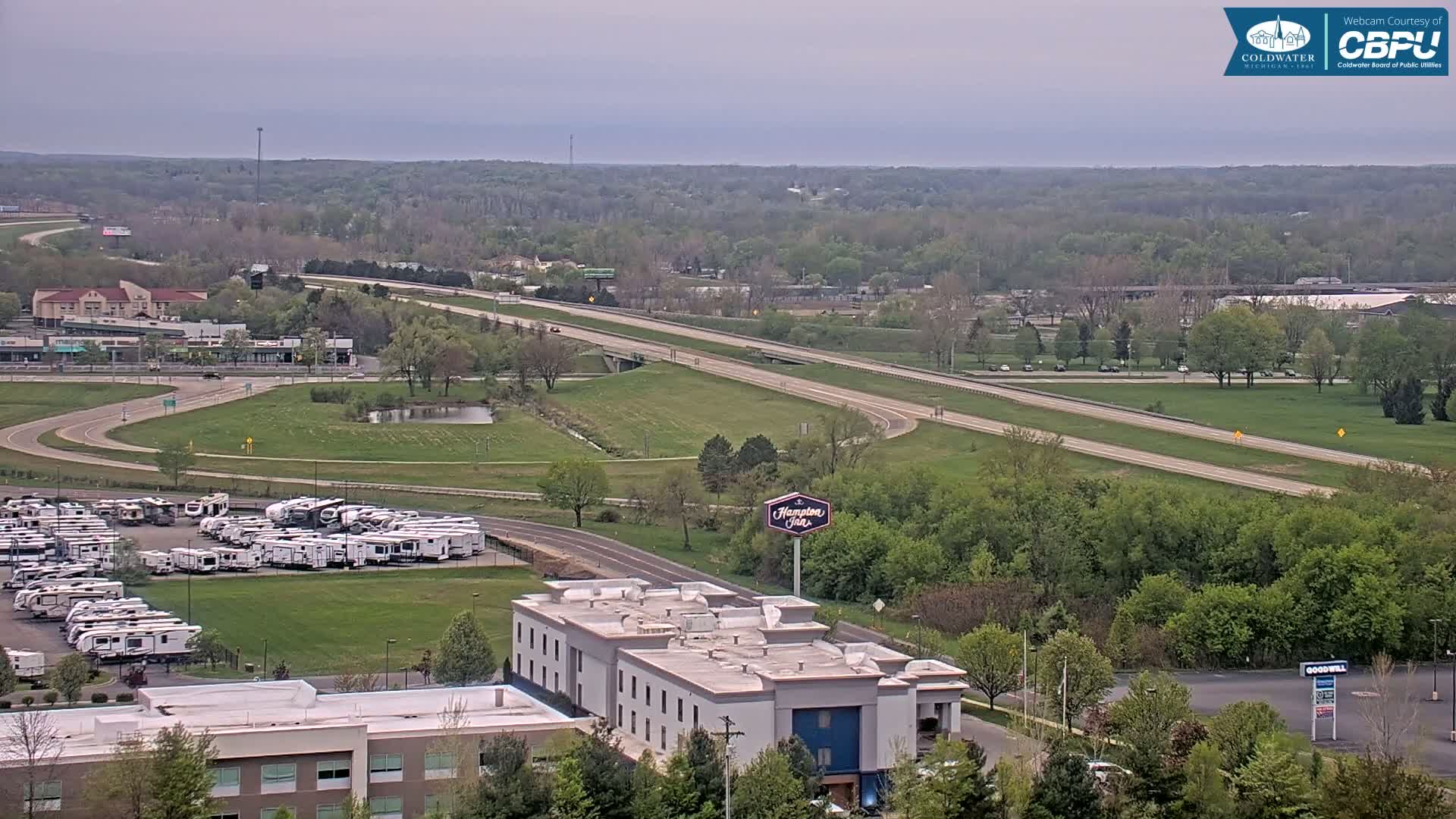 A highway stretches across a verdant landscape under an overcast sky, with a hotel and a large RV park visible nearby.