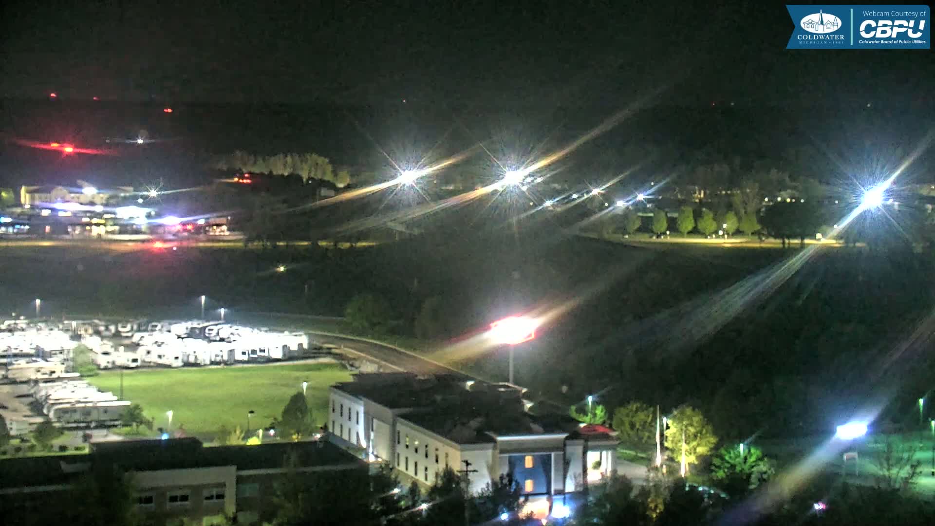 A nighttime view of a landscape featuring a parking lot filled with RVs, a hotel, and distant lights, under clear skies.