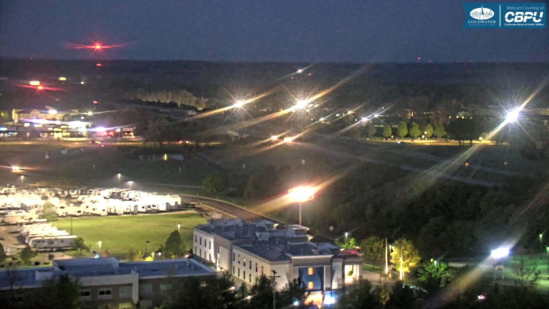 A nighttime aerial view of a town shows a highway with car lights streaking, a large RV park, and a multi-story building.