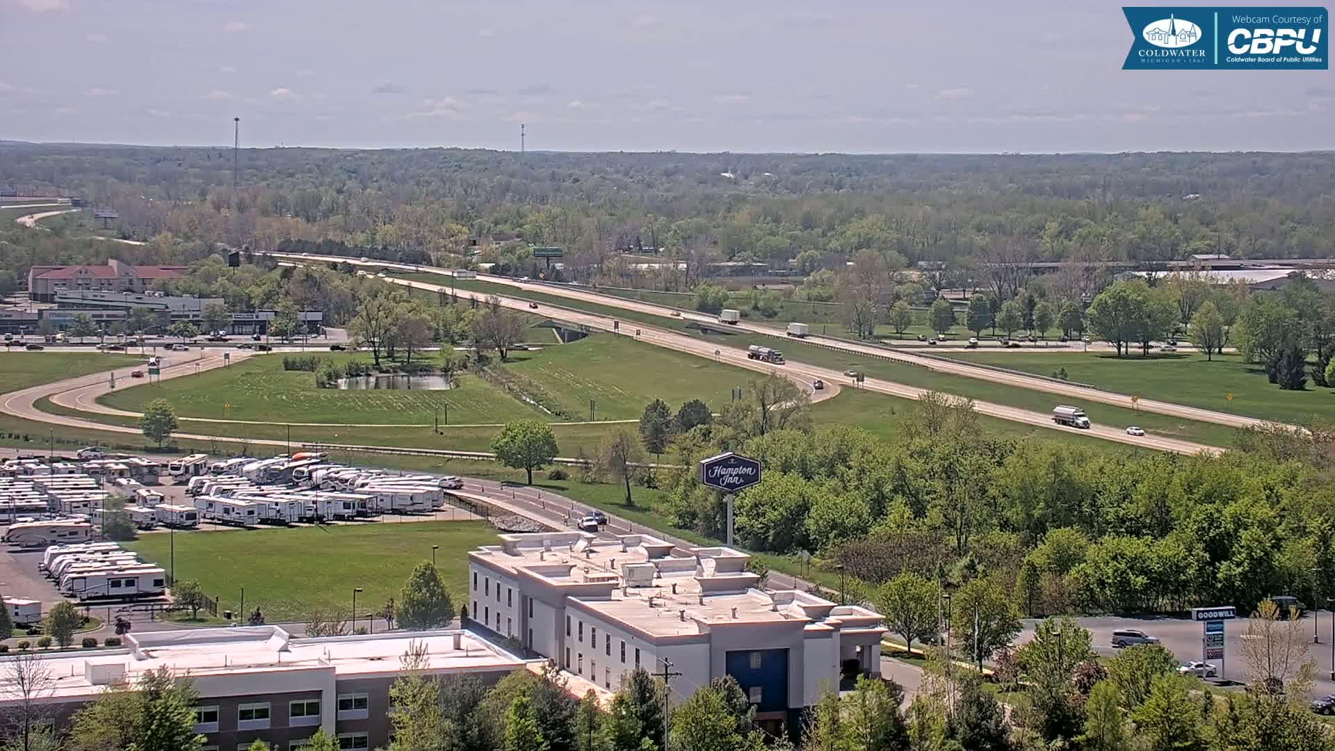 A highway cuts through a verdant landscape under a partly cloudy sky, with a large RV park and a Hampton Inn visible below, alongside some other buildings and a Goodwill store.