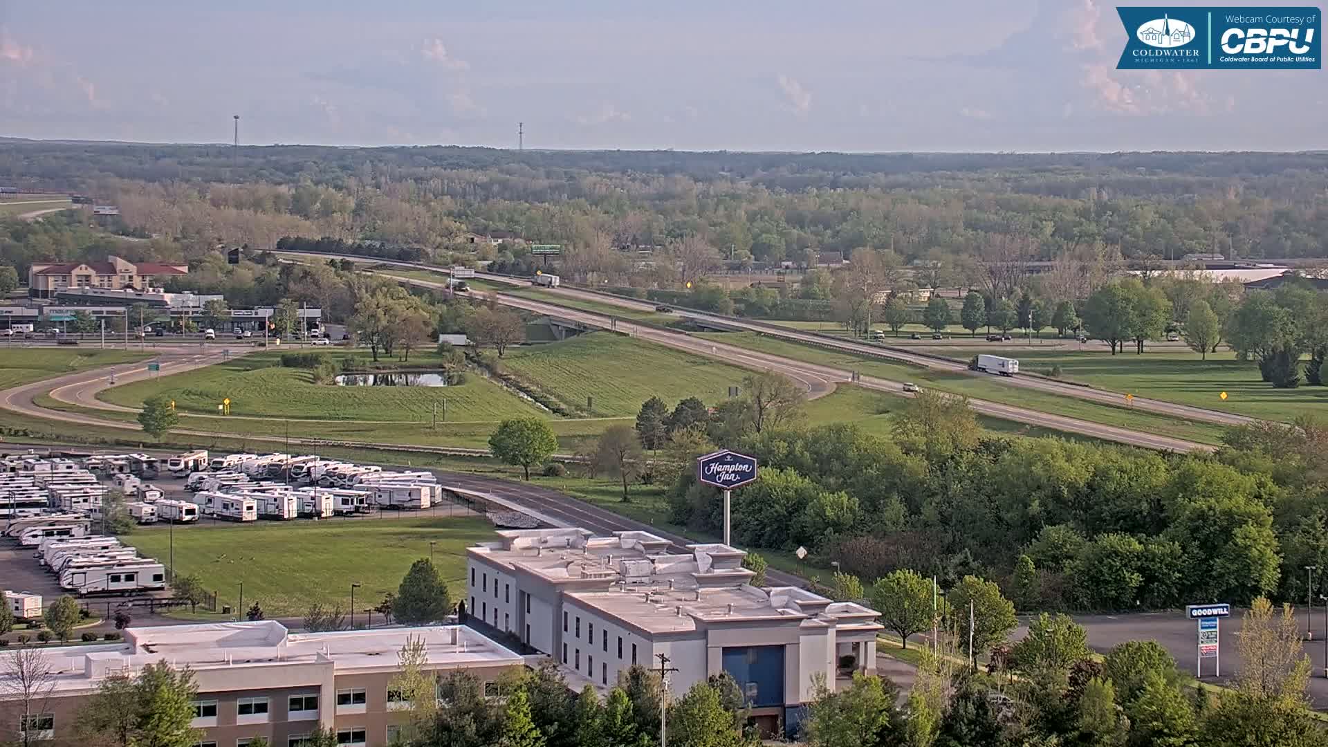 An aerial view shows a highway, a Hampton Inn, a large RV parking lot, and green, leafy trees under a partly cloudy sky.