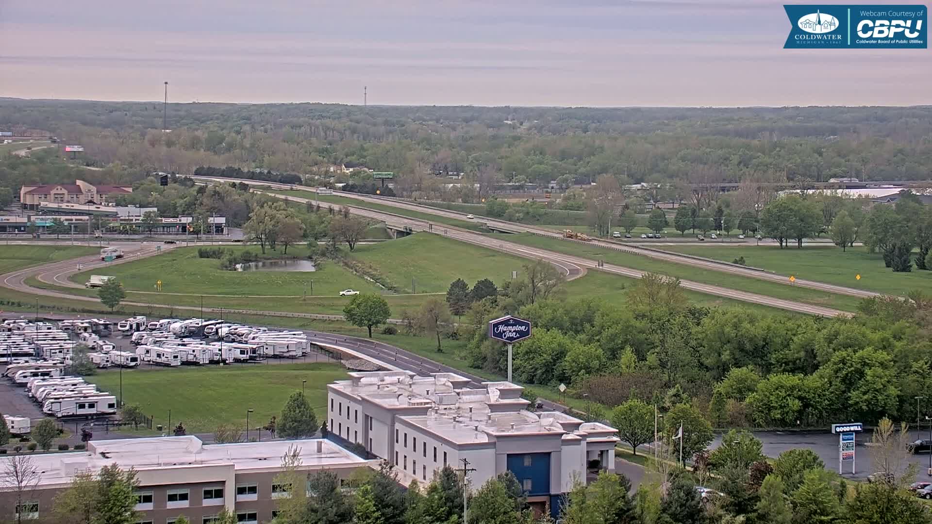 A sprawling landscape of green trees and a highway curves past a Hampton Inn and a large RV park under an overcast sky.
