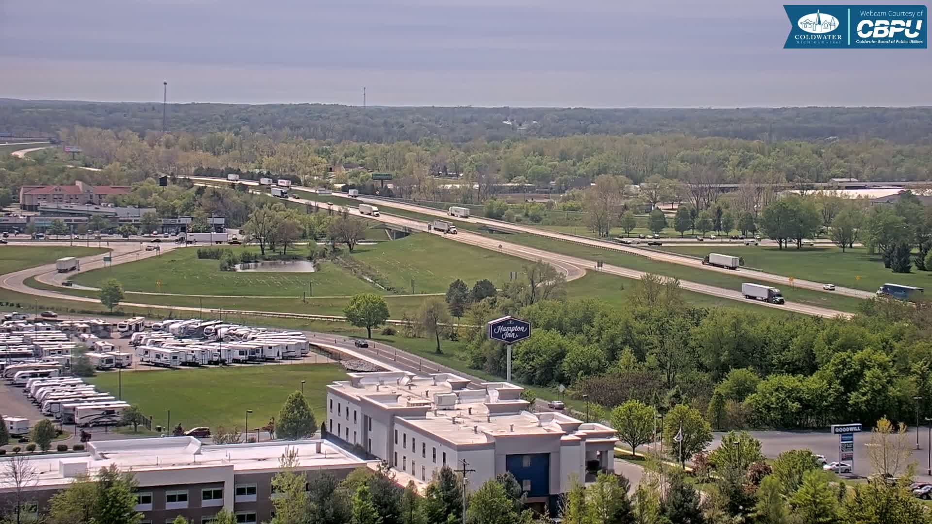 An aerial view shows a highway with several semi-trucks driving on it, a large RV park, a Hampton Inn, and surrounding green trees under a partly cloudy sky.