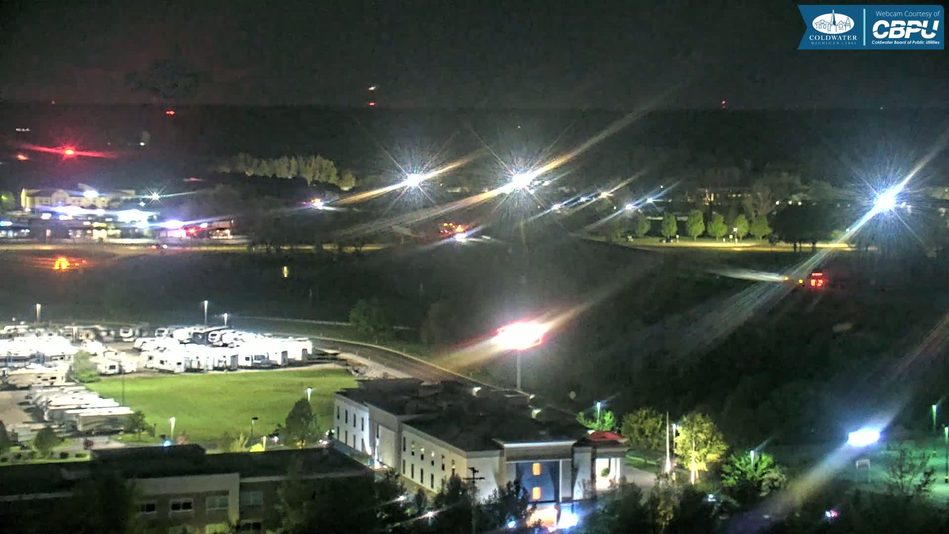 A nighttime aerial view shows a highway with bright lights, a parking lot full of RVs, and a multi-story building, all surrounded by dark green trees and grass.