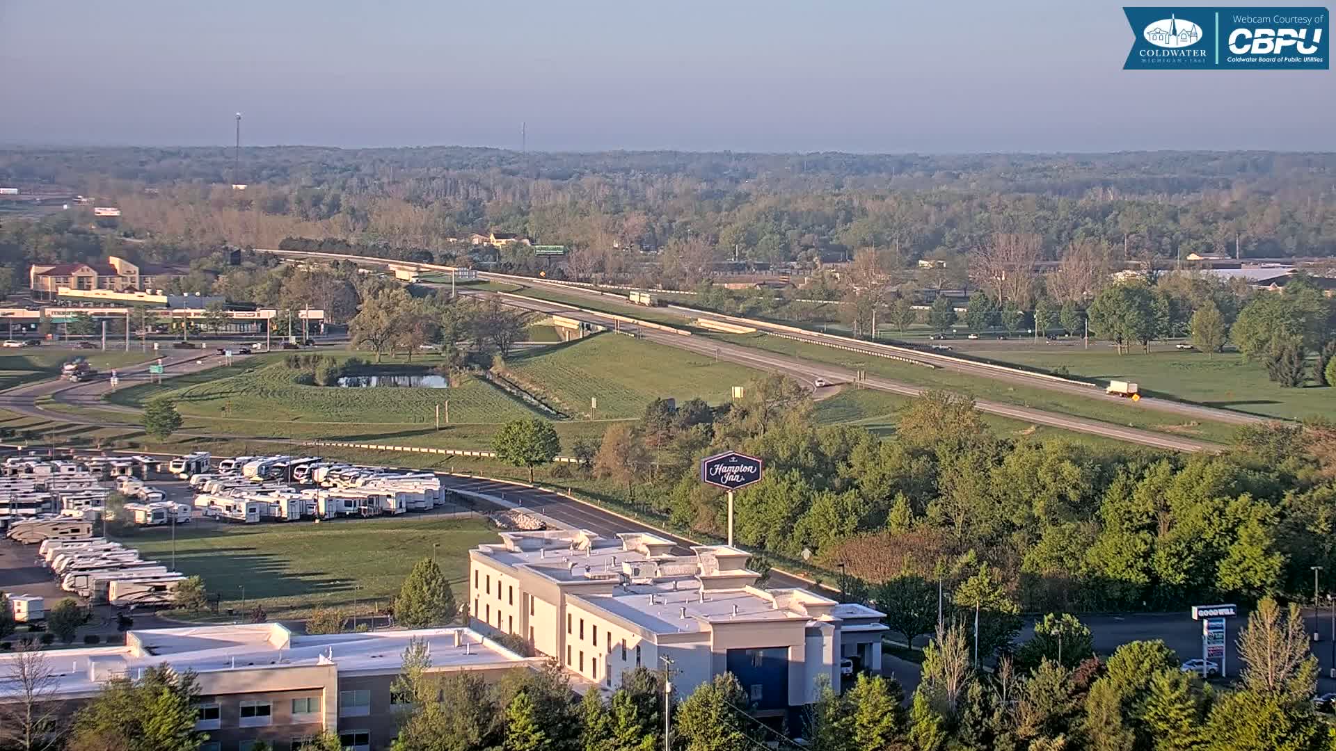 A sprawling landscape featuring a highway, a large RV park, a Hampton Inn, and a mix of green trees and grassy areas under a hazy, clear sky.