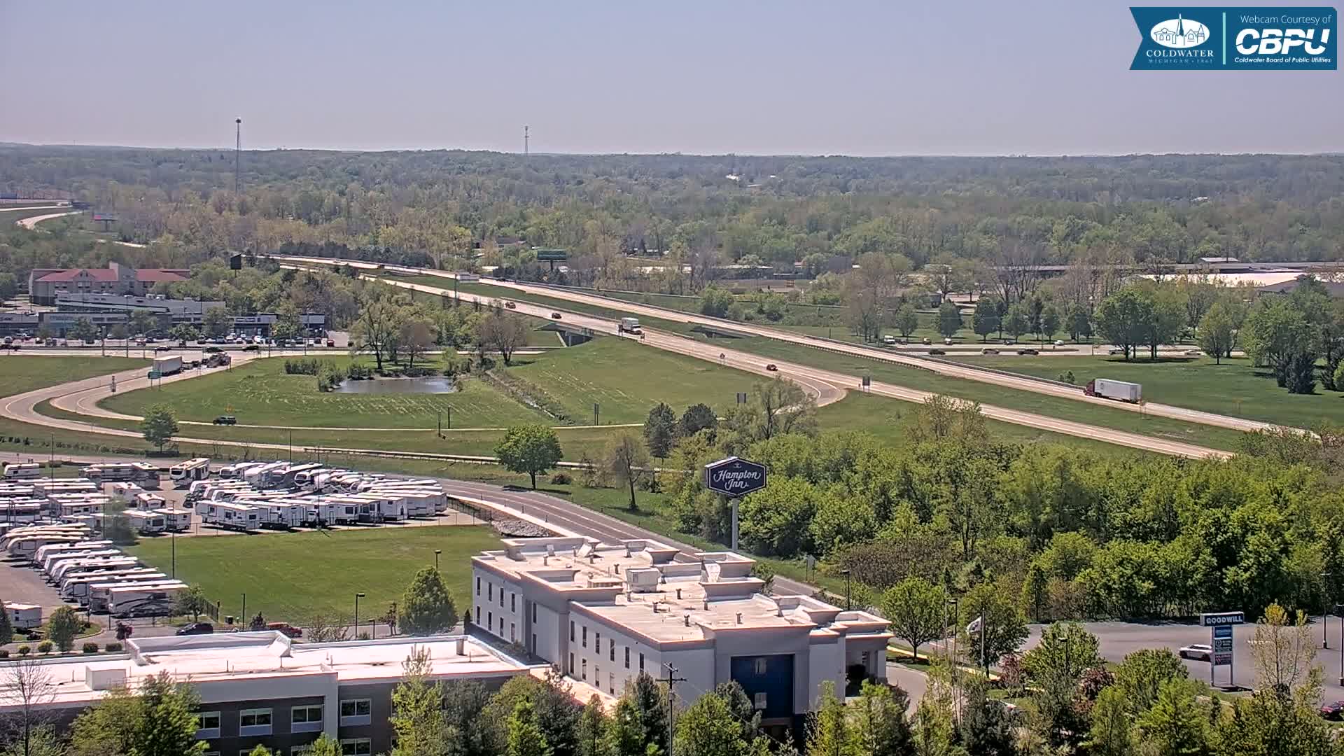 A highway with light traffic cuts through a green landscape dotted with buildings, including a hotel and a large RV park, under a clear, sunny sky.