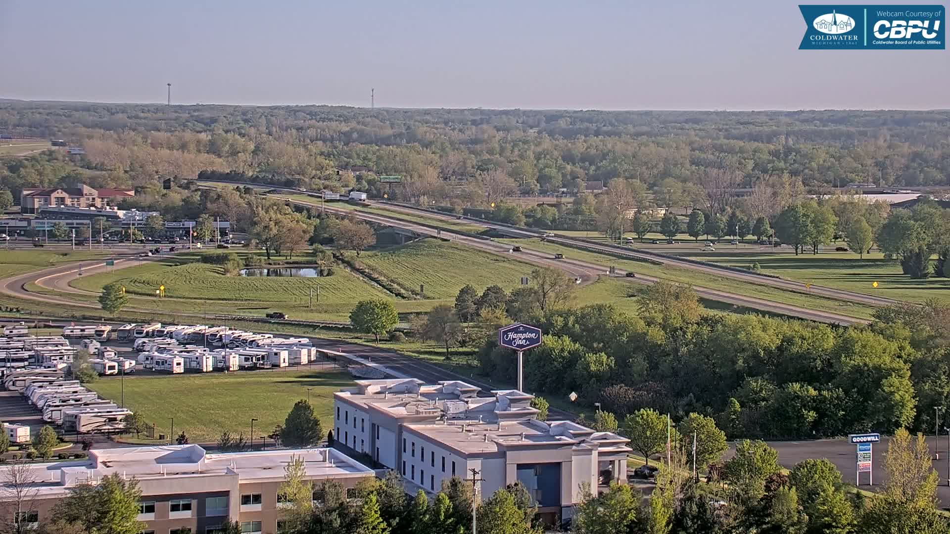 A high-angle, long shot shows a highway with few cars, a Hampton Inn, a large RV park, and a surrounding landscape of green trees and grassy fields under a clear, bright sky.