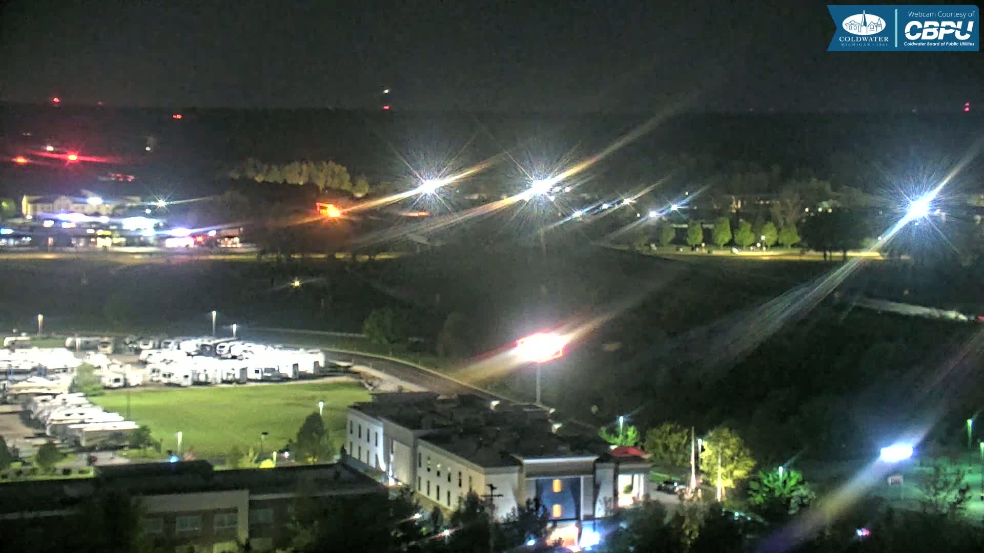 A nighttime aerial view shows a parking lot full of RVs, a multi-story building, and roadways with car lights streaking across the frame.