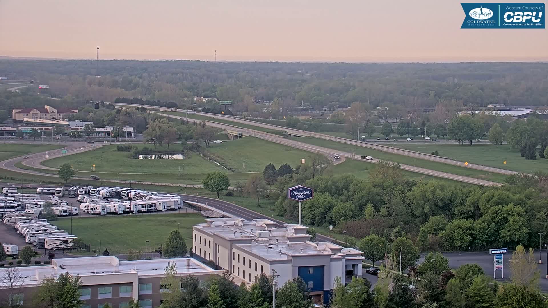 A sprawling landscape at dawn features a multi-lane highway, a large parking lot filled with recreational vehicles, a hotel, and surrounding green spaces.
