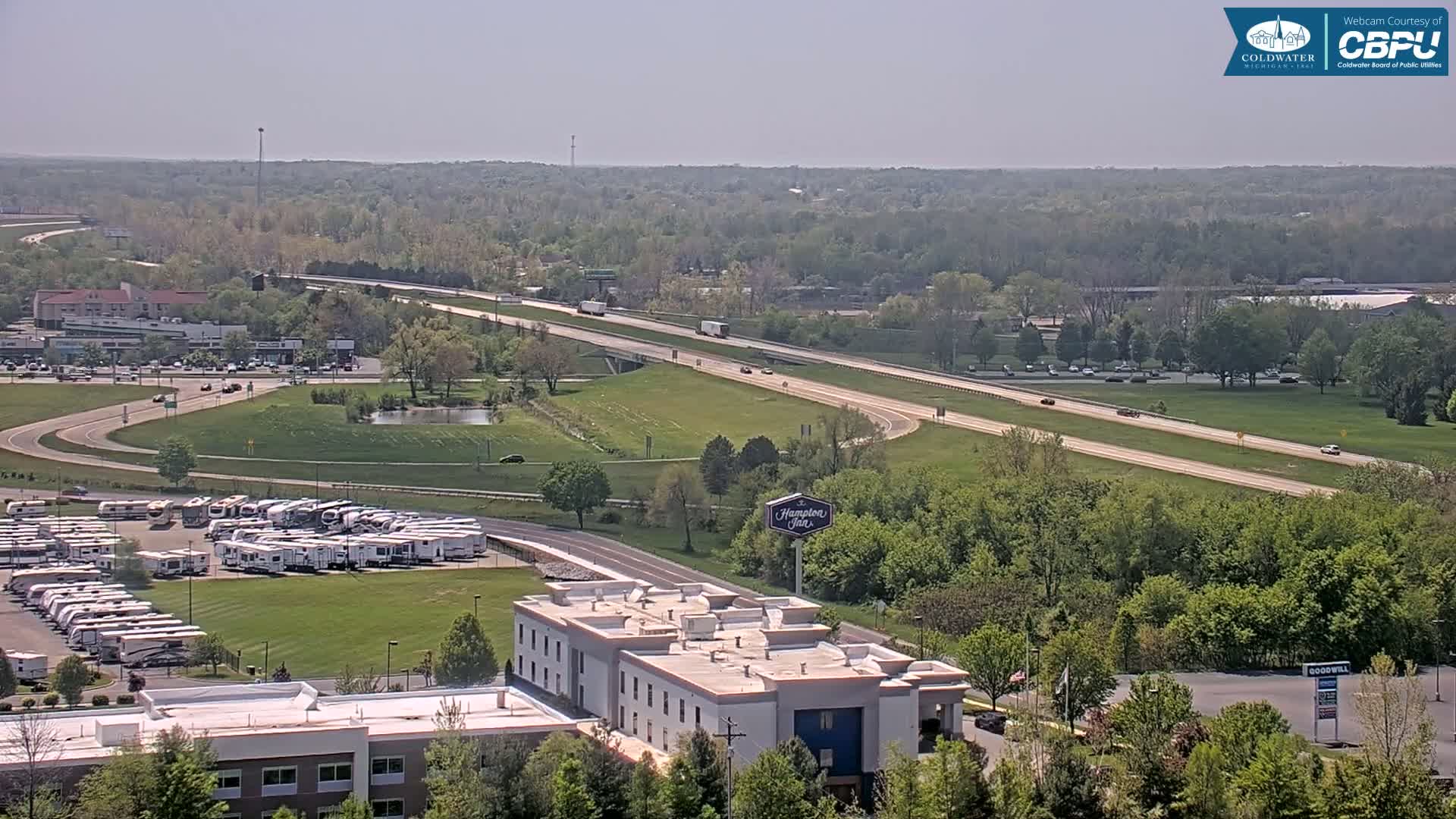 A highway with moderate traffic runs through a green landscape dotted with buildings, including a hotel and a large RV park, under a hazy sunny sky.