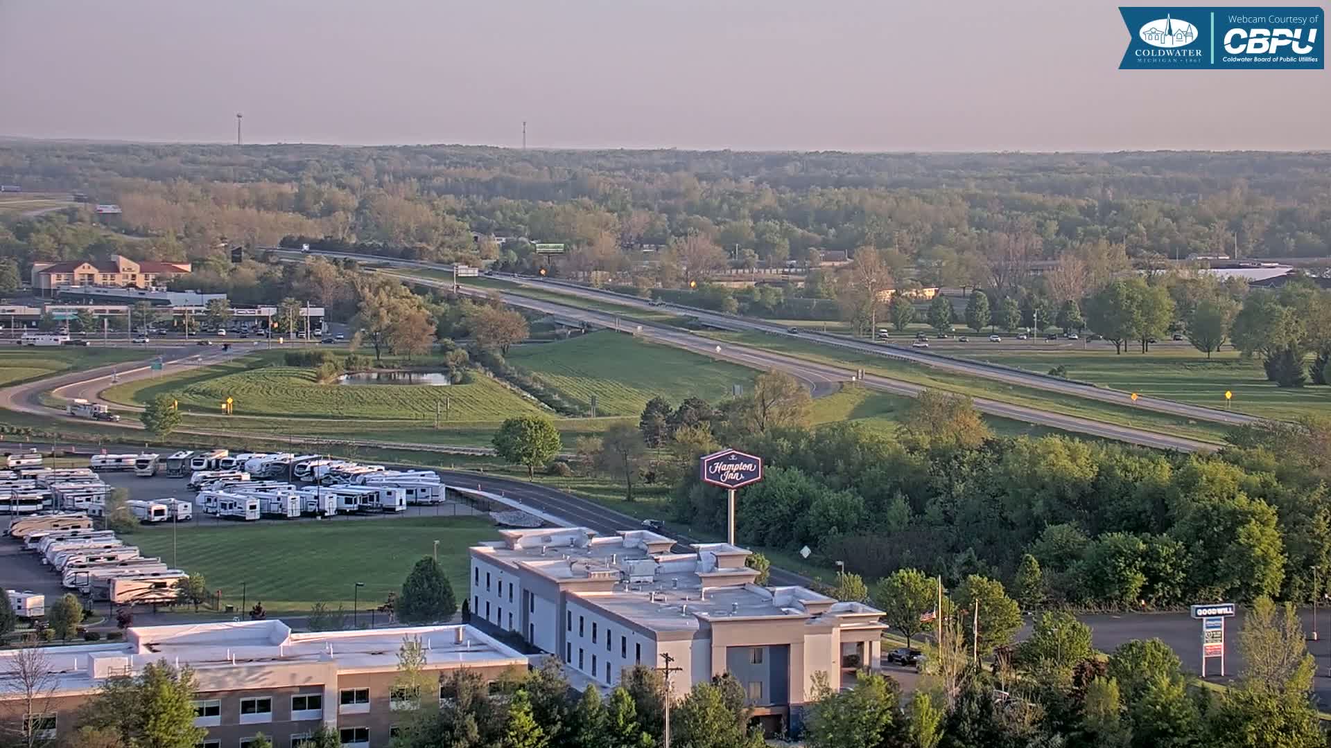 A high-angle view of a highway intersection with a Hampton Inn, a large RV park, and surrounding green trees under a hazy sky.