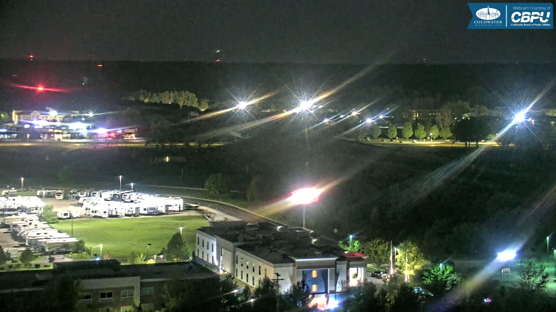A nighttime aerial view shows a motel, a large RV park, and a wooded area illuminated by streetlights and vehicle lights under a clear, dark sky.