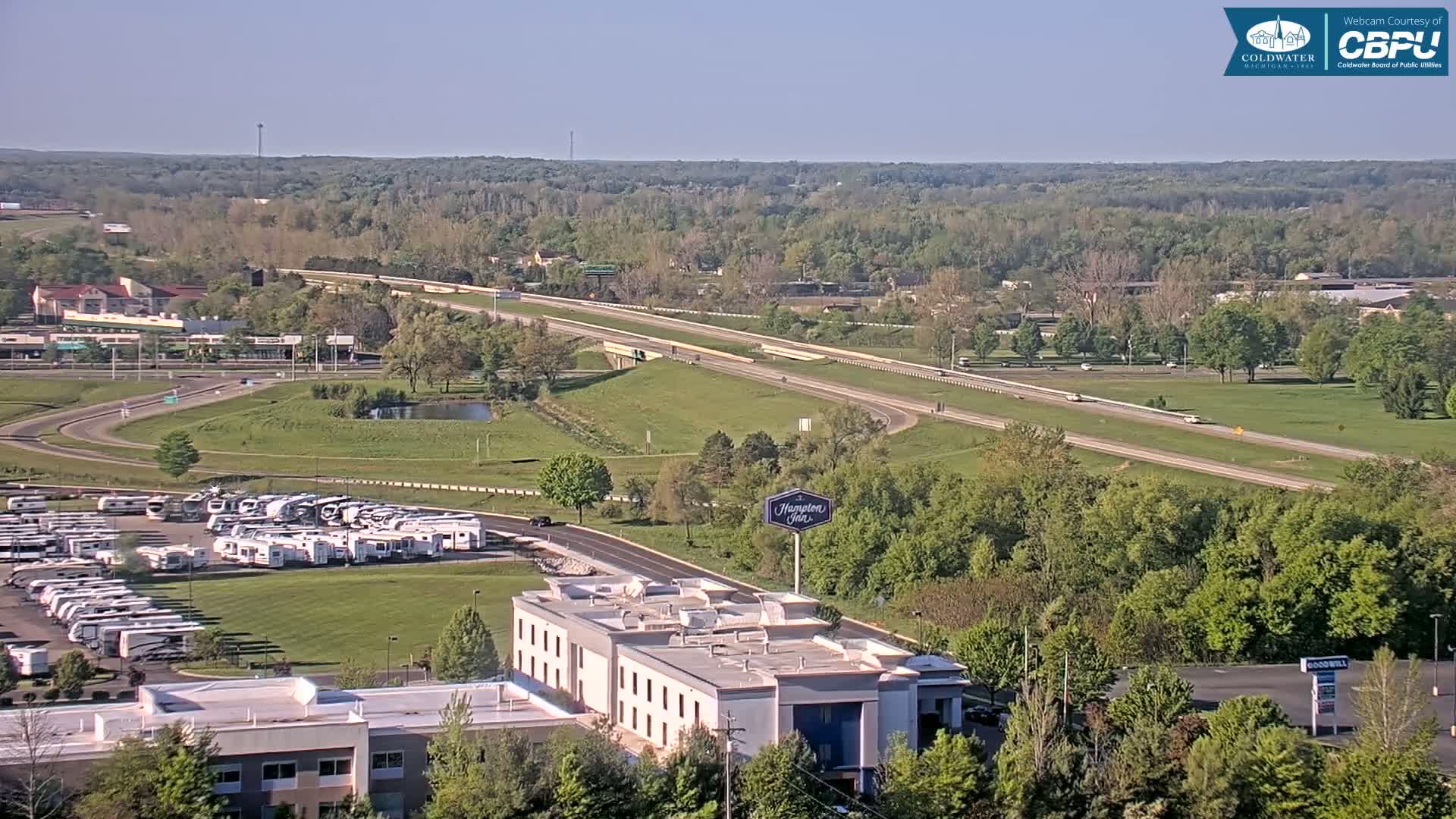 A long highway cuts through a verdant landscape under a clear sky, with a large parking lot filled with RVs and a hotel visible nearby.