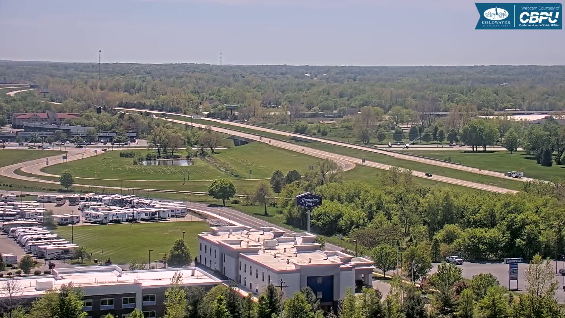 A high-angle, long shot on a sunny day shows a highway and overpass cutting through a green landscape dotted with buildings, including a Hampton Inn, a large RV lot, and other commercial structures.