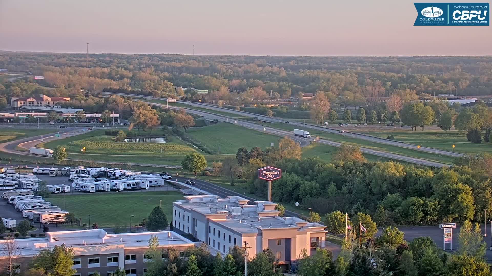 An elevated view of a highway and surrounding area, including a hotel, RV park, and green spaces, under a clear, light sky at sunset.